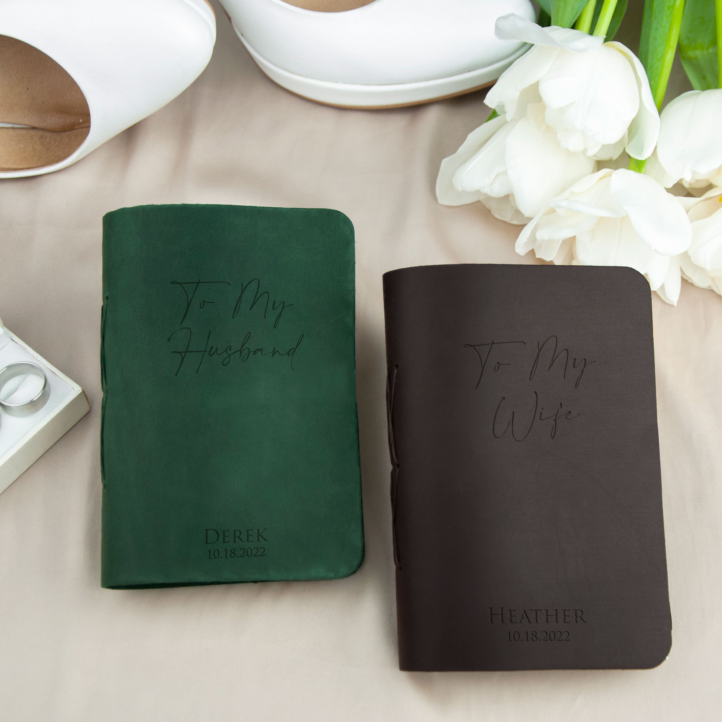 two personalized notebooks, one green and one brown, with the names "To My Husband" and "To My Wife" printed on them. The notebooks are placed on a table with a pair of white shoes and a bouquet of white flowers.