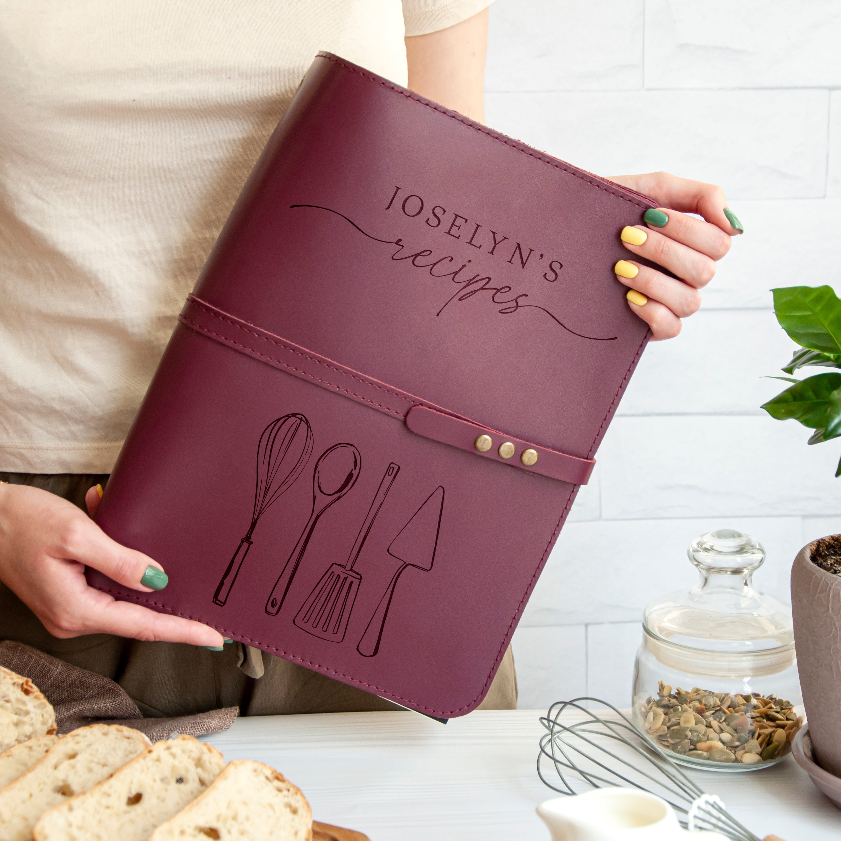 A person is holding a burgundy leather recipe book with the title "Joslyn's recipes" printed on it. The book is decorated with illustrations of kitchen utensils, such as a whisk and a spatula. The person is standing next to a table with various food items, including bread and a glass jar.