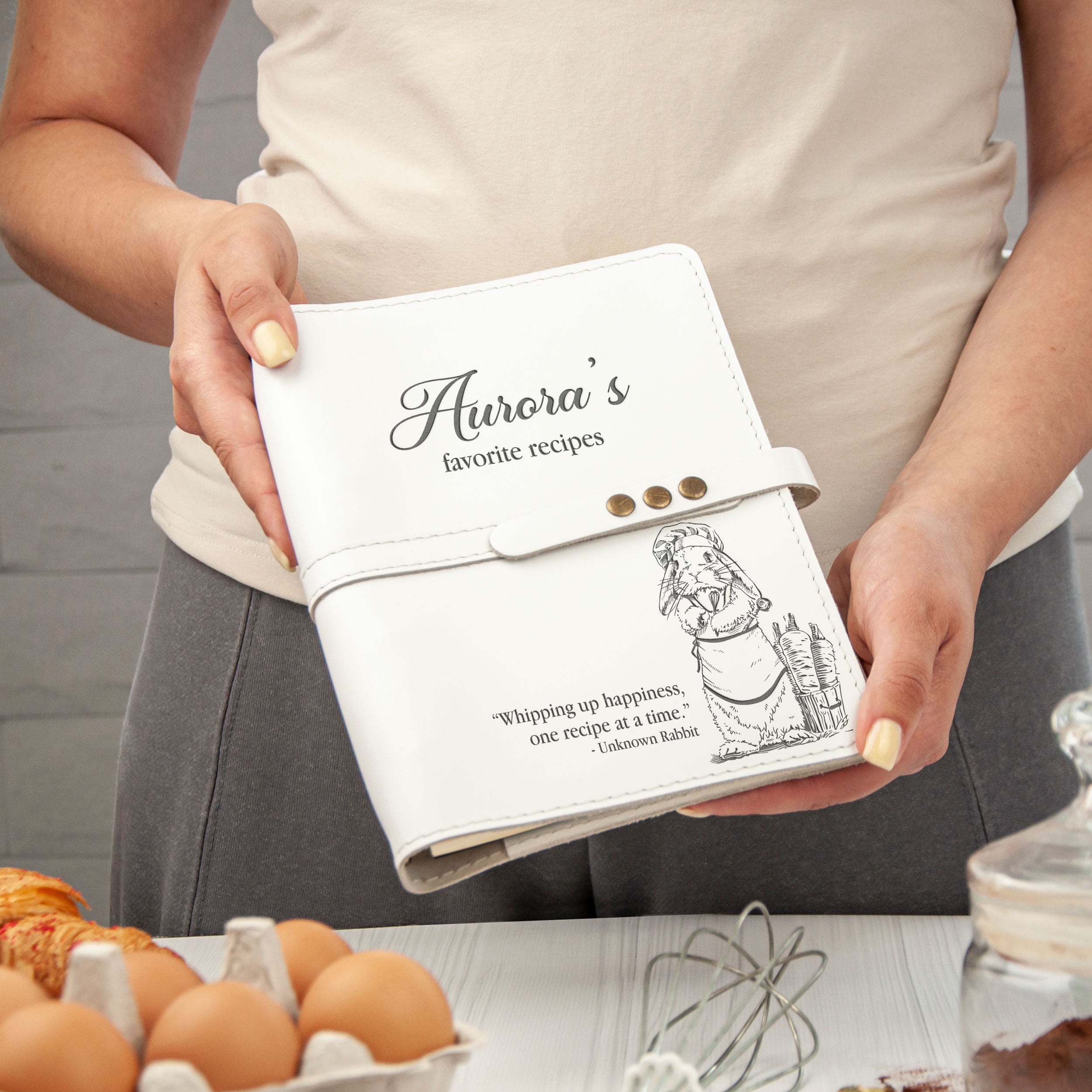 A person is holding a white leather-bound recipe book with a personalized cover featuring a cartoon character and the text "Aurora's favorite recipes".