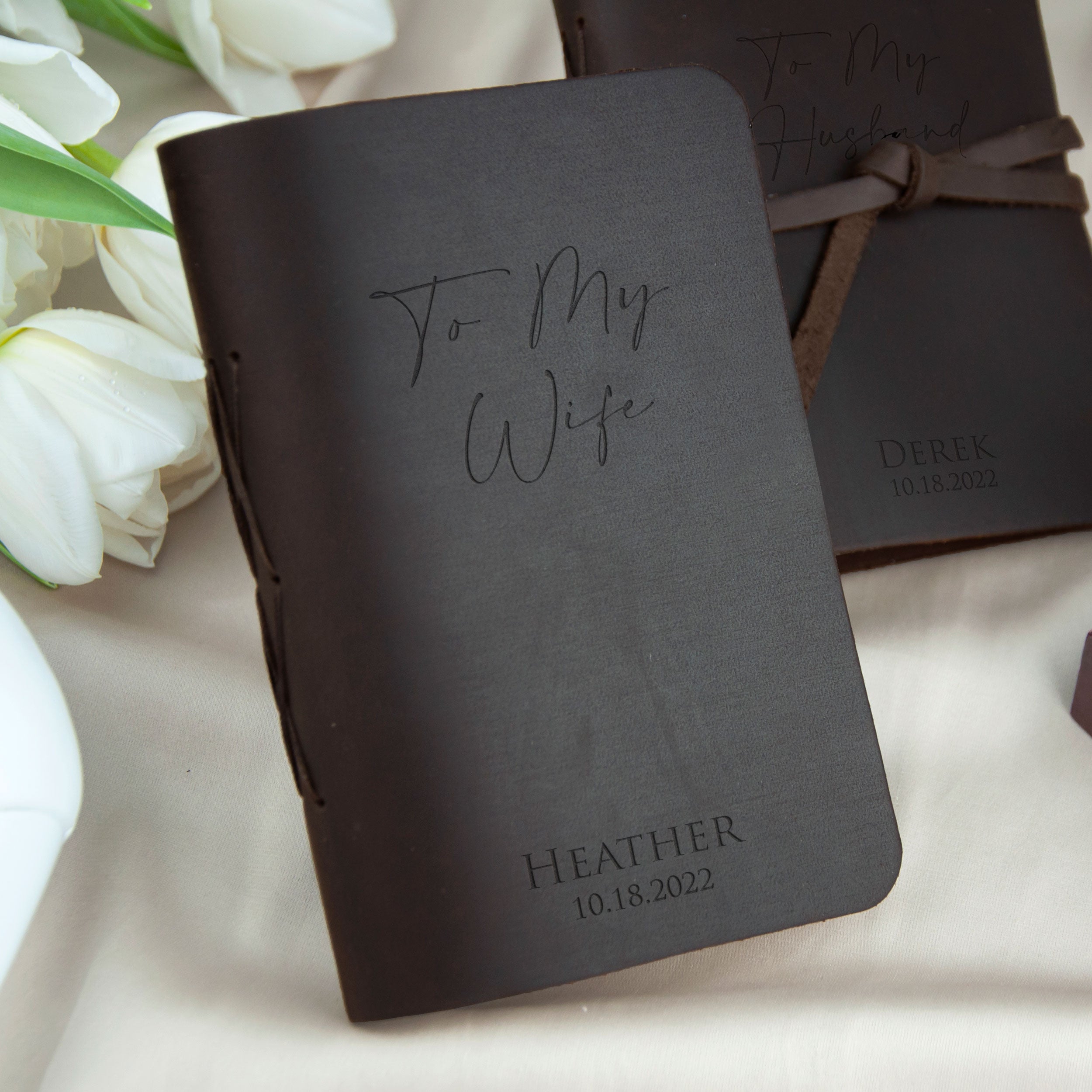 A black leather-bound book titled "To My Wife" sits on a white surface, with a bouquet of white flowers in the background.