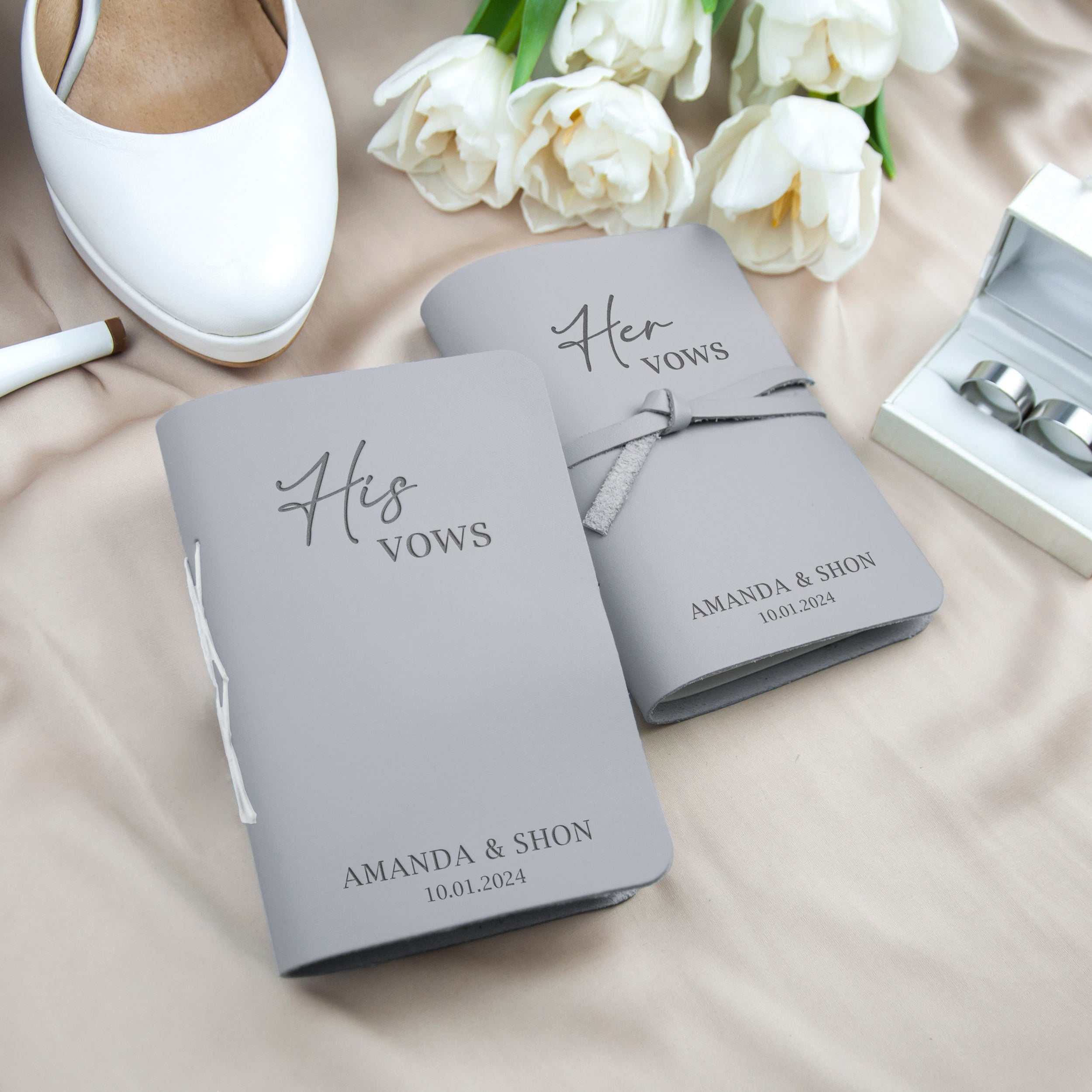 two grey books with the text "His Vows" and "Her Vows" printed on them, along with the names "AMANDA & SHON" and the date "10.01.2024". The books are placed on a beige surface, with a pair of white shoes and a bouquet of white flowers in the background.