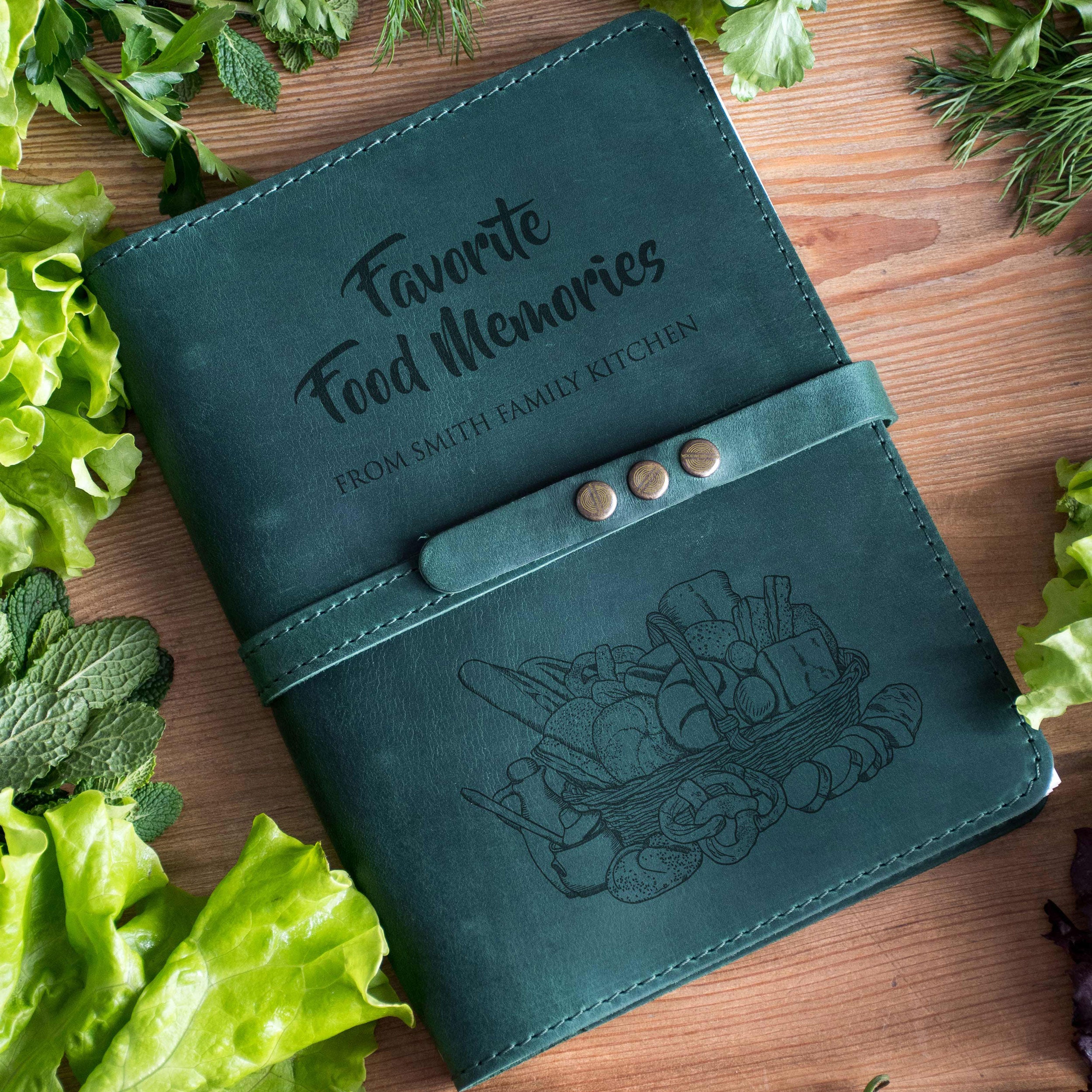 A green leather-bound notebook titled "Favorites Food Memories" with a picture of a basket of food on the cover, surrounded by fresh green vegetables on a wooden surface.