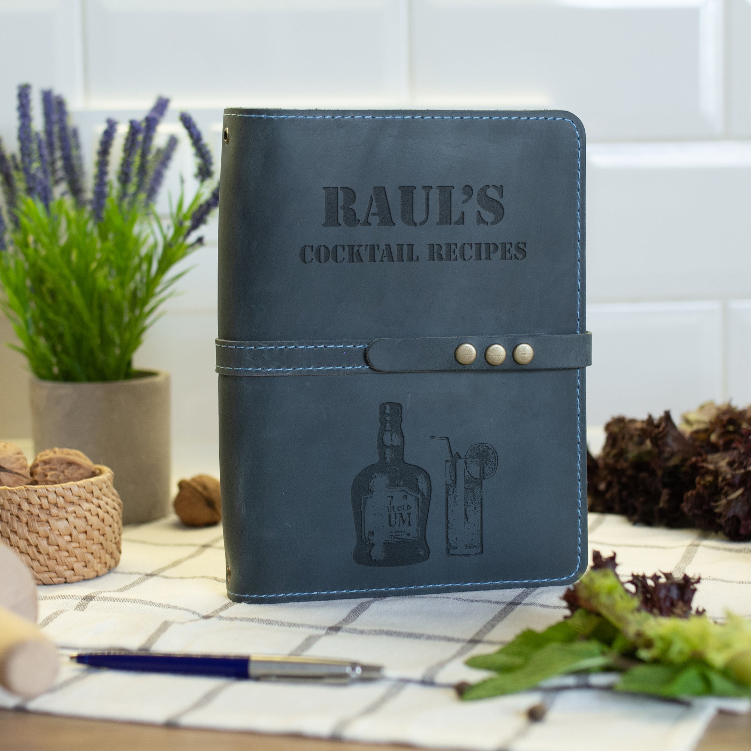 A leather-bound cocktail recipe book with the title "RAUL'S COCKTAIL RECIPES" printed on the cover, placed on a table with various items such as a potted plant, a pen, and some food.