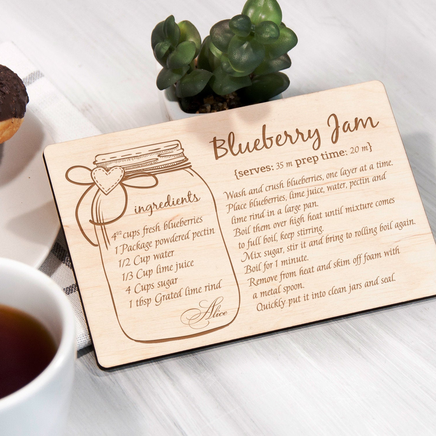 A wooden recipe card with a blueberry jam recipe, placed on a table next to a cup of tea and a potted plant.