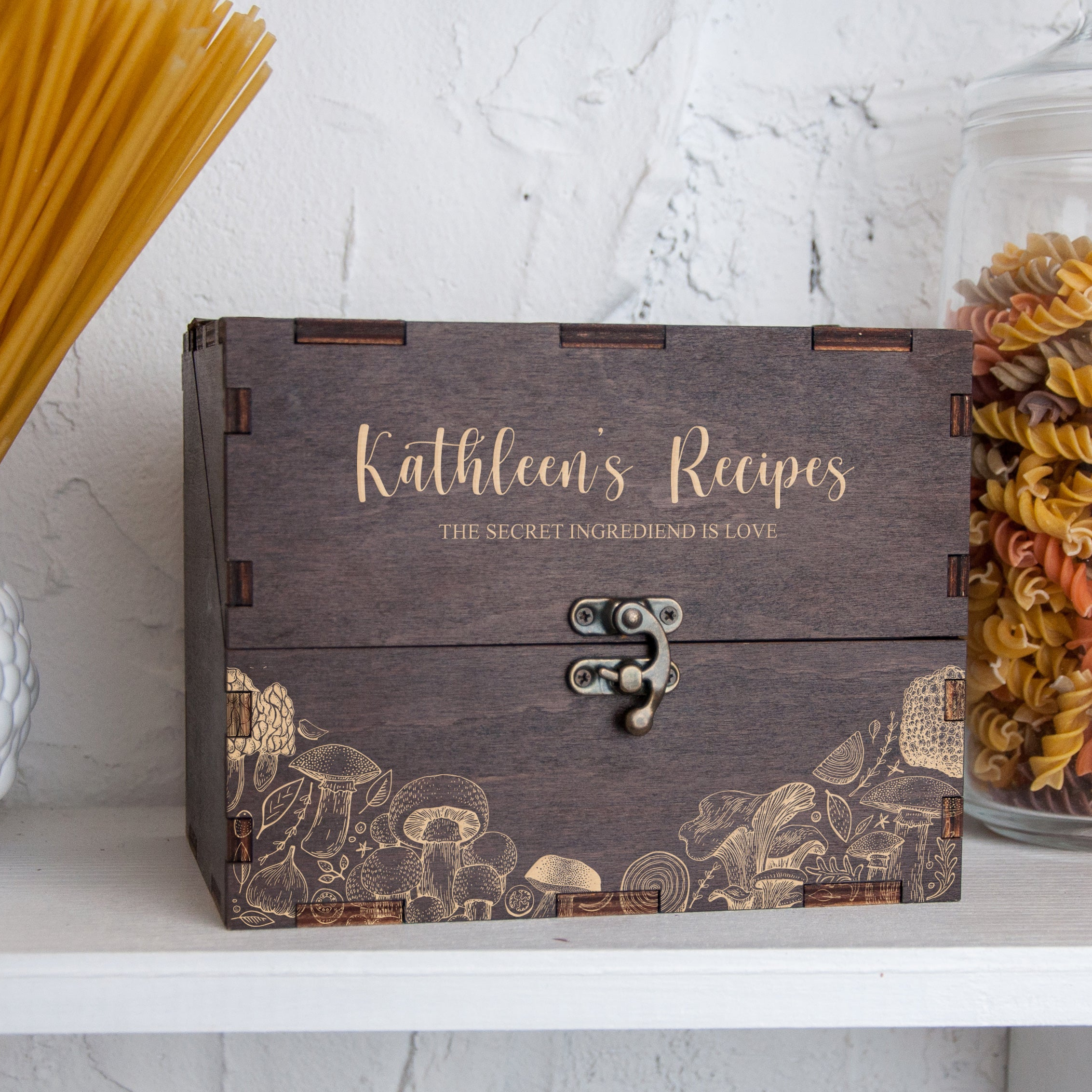 A wooden box with the name "Kathleen's Recipes" printed on it, sitting on a shelf next to a glass jar filled with pasta.