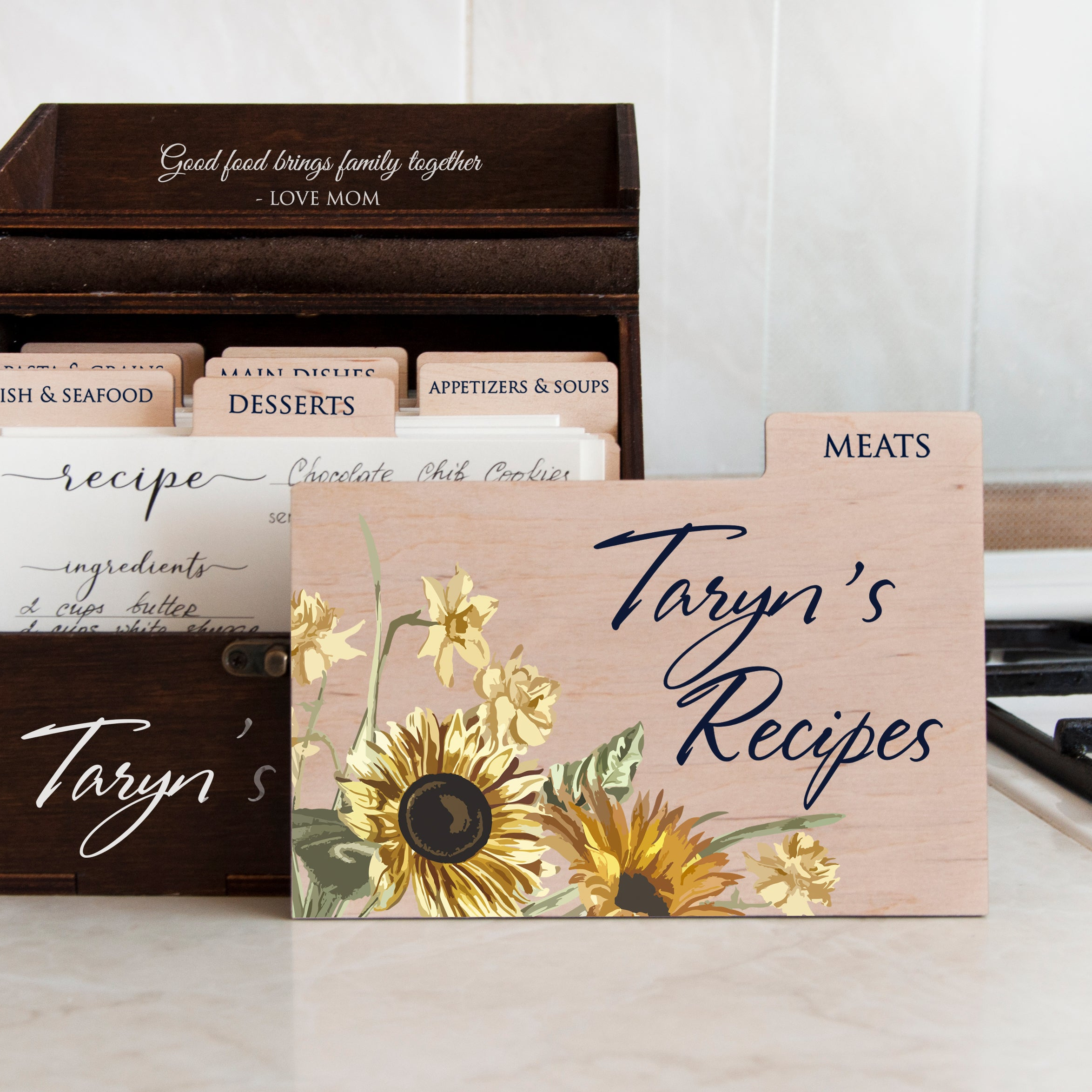 a wooden recipe box with a floral design, displaying the name "Taryn's Recipes" and a list of ingredients. The box is placed on a countertop, with a wooden tray containing more recipe cards visible in the background.
