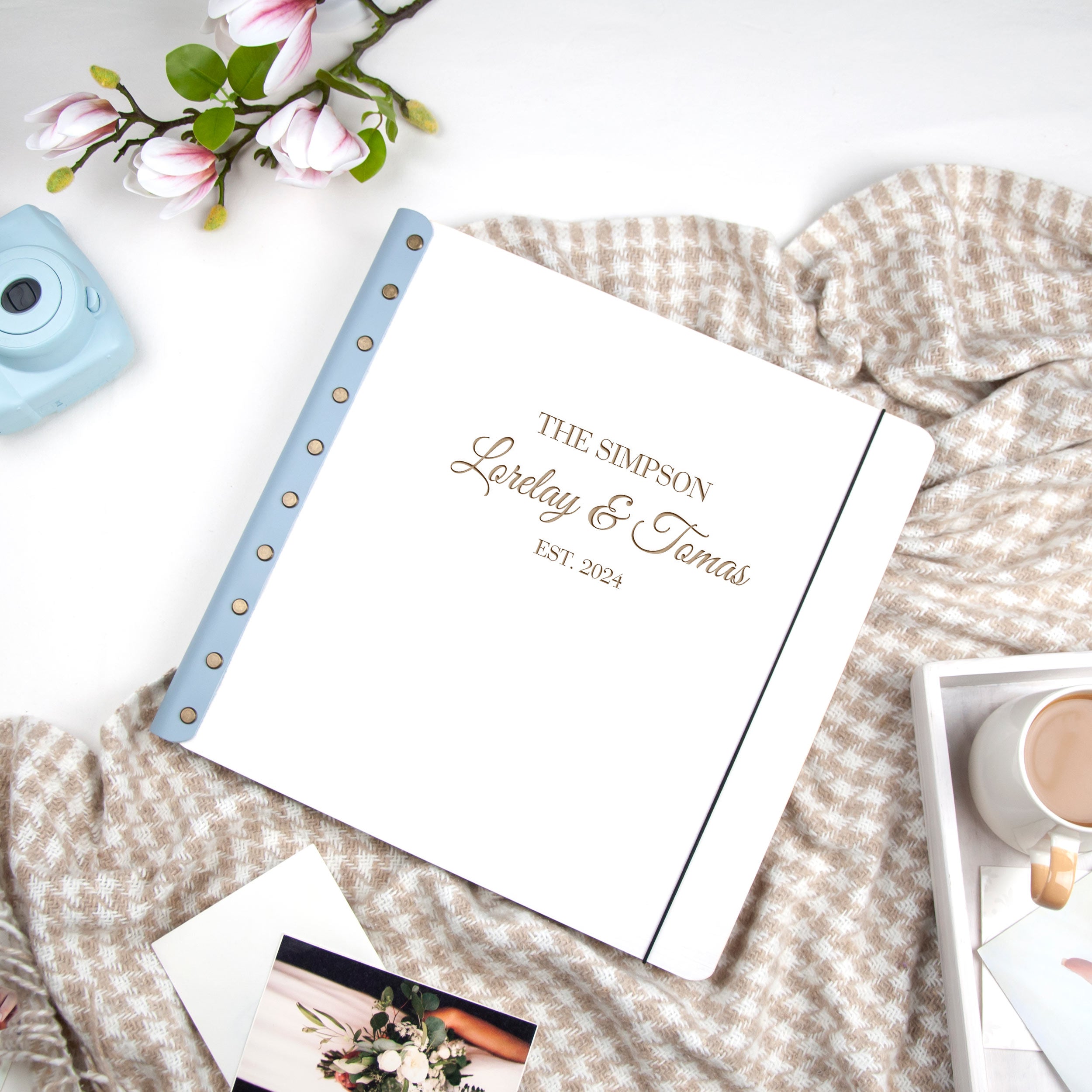 a white notebook with the name "The Simpson" printed on it, placed on a table with a floral arrangement, a camera, and a mug of coffee.