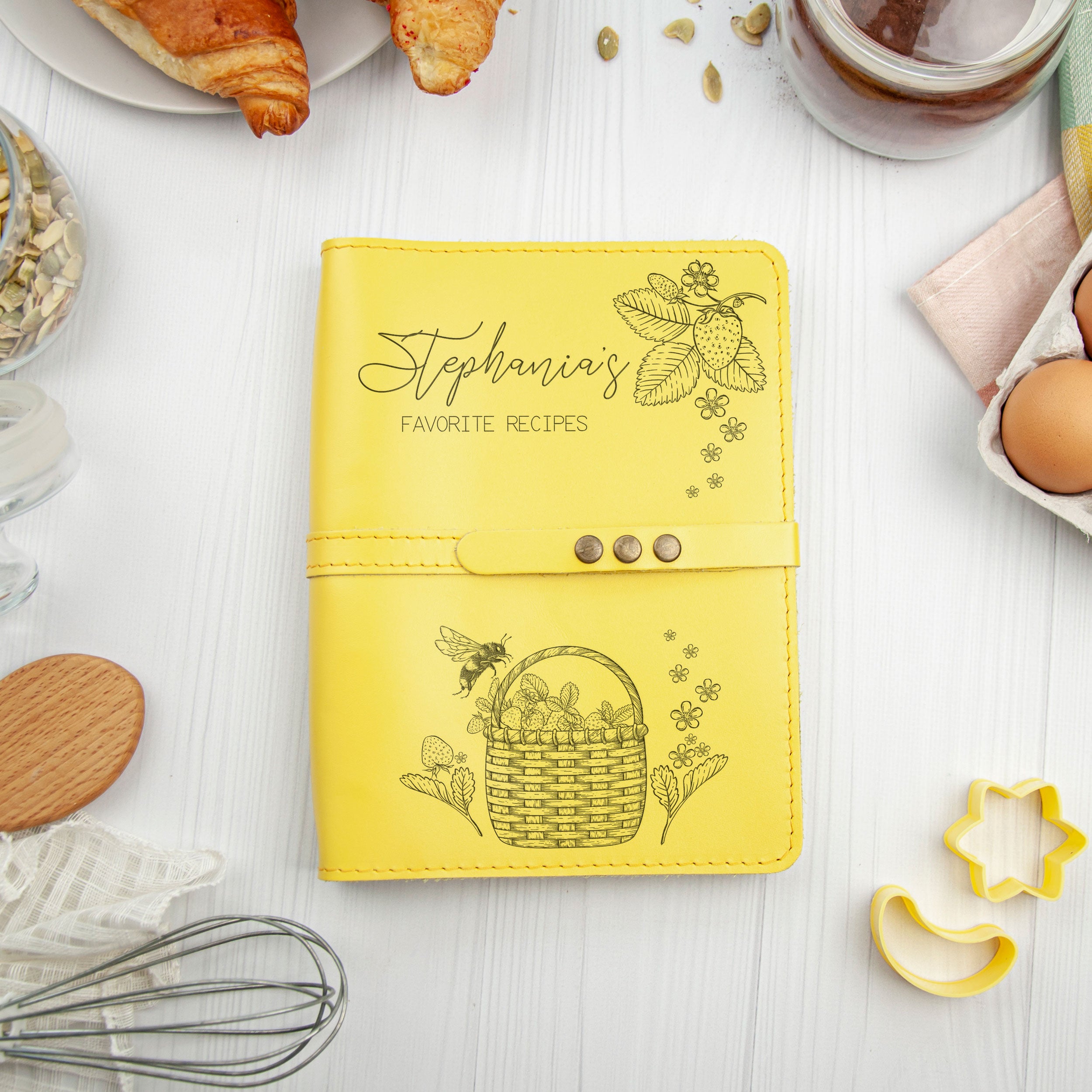 a yellow recipe book with a basket of fruit and a bee on the cover, surrounded by various kitchen items such as a croissant, a glass jar, and a wooden spoon.