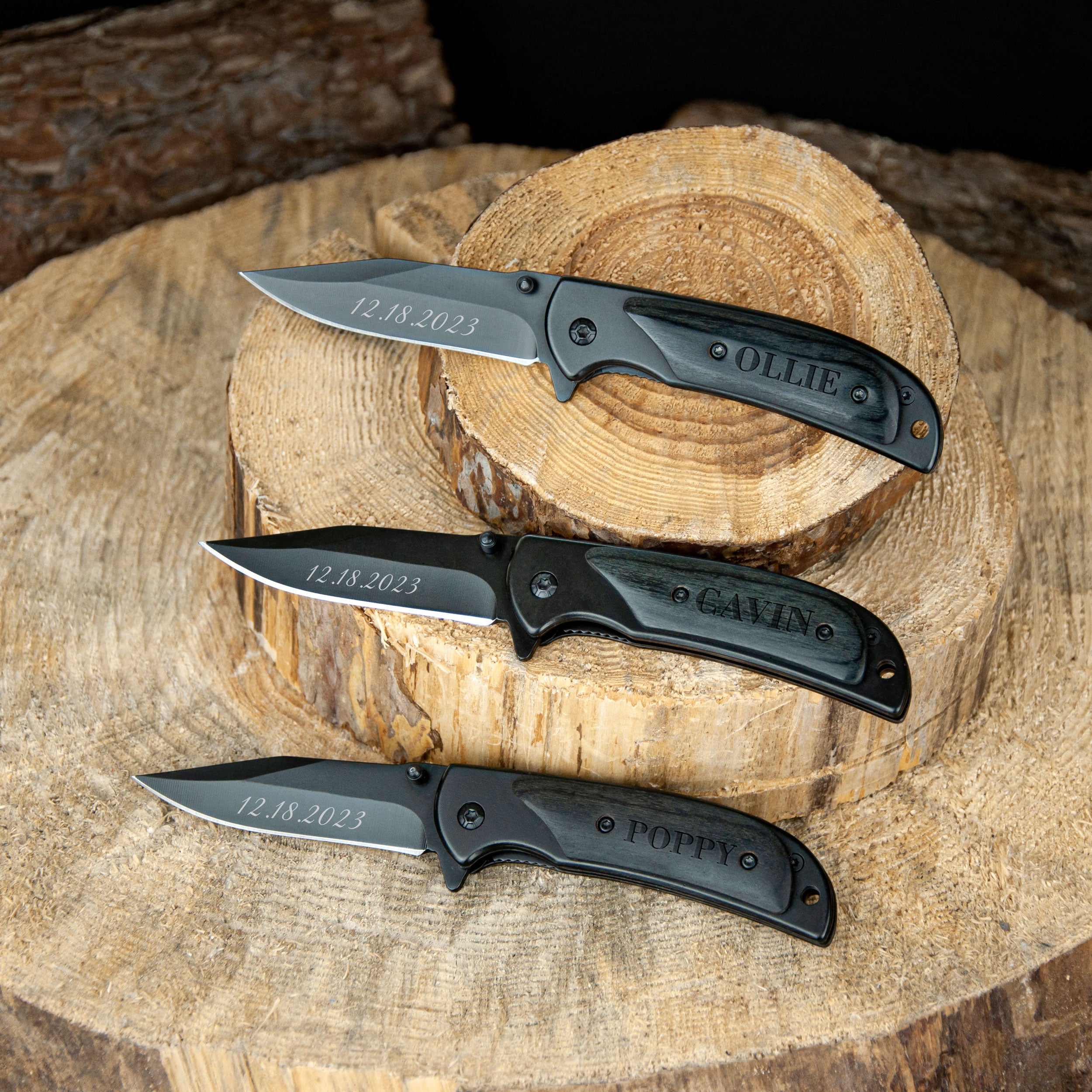 Three black pocket knives with personalized engravings are displayed on a wooden stump.