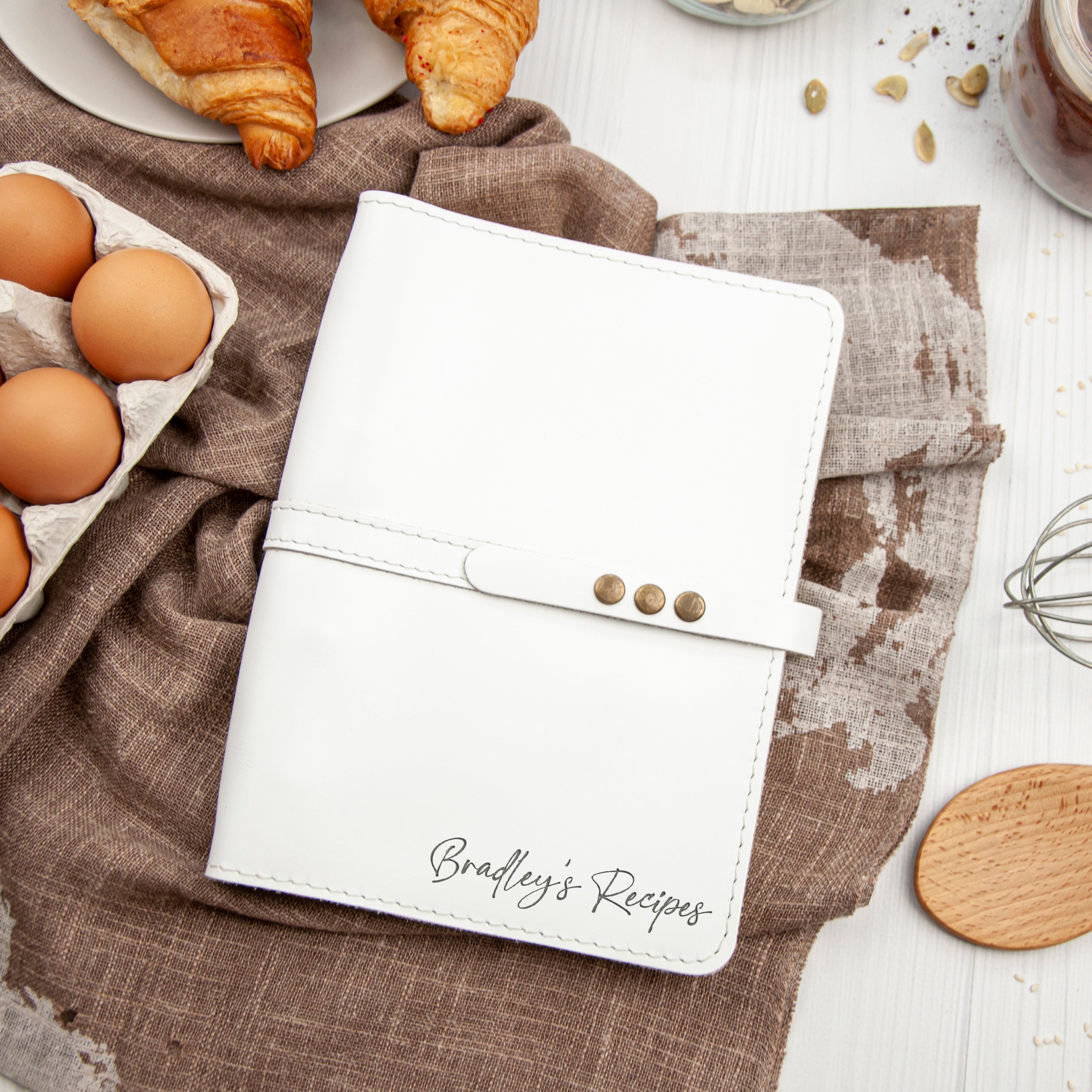 A white recipe book with the name "Bradley's Recipes" printed on it, placed on a table with various food items such as eggs, a croissant, and a wooden spoon.