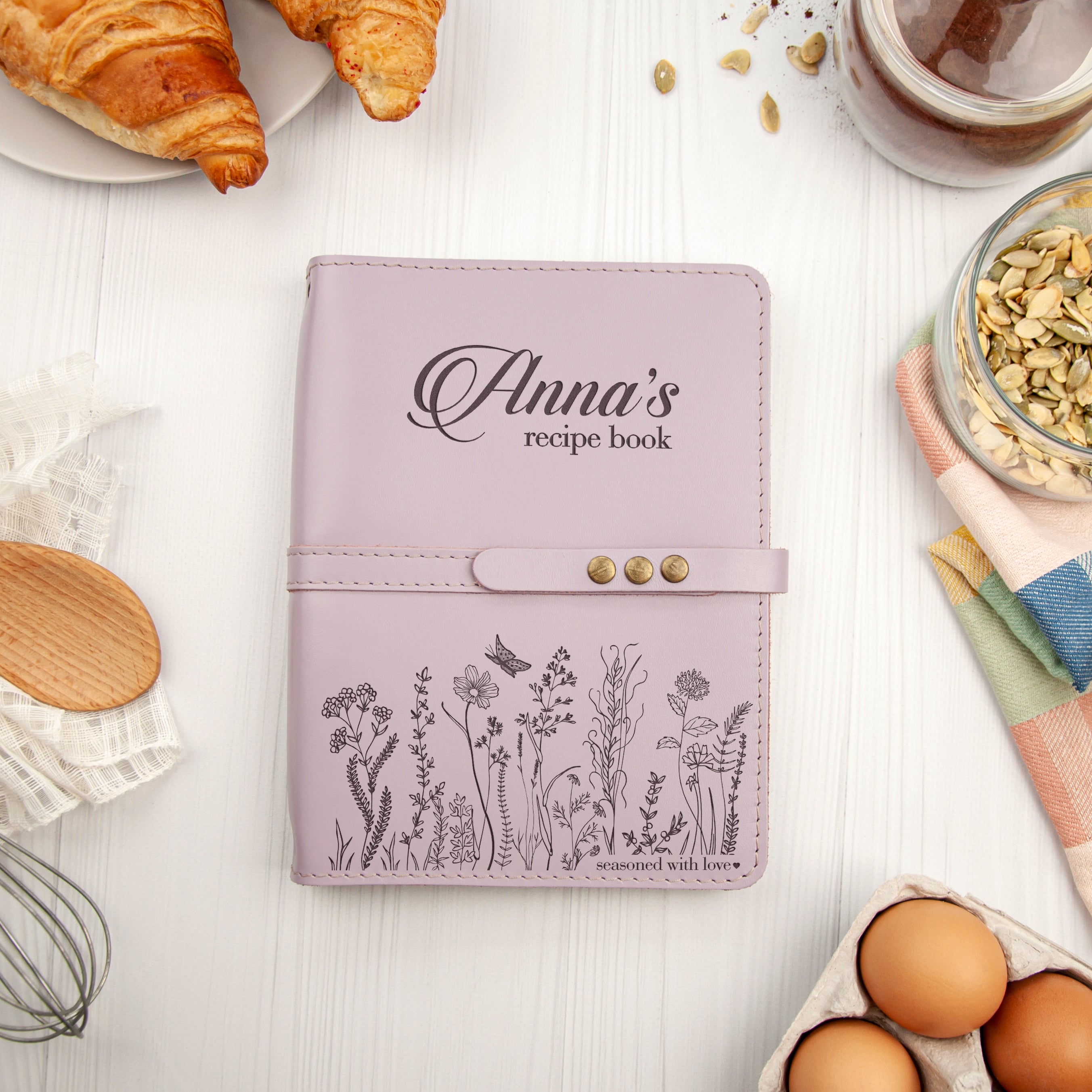 a pink recipe book with a floral design, surrounded by various kitchen items such as a croissant, a bowl of nuts, and a carton of eggs.