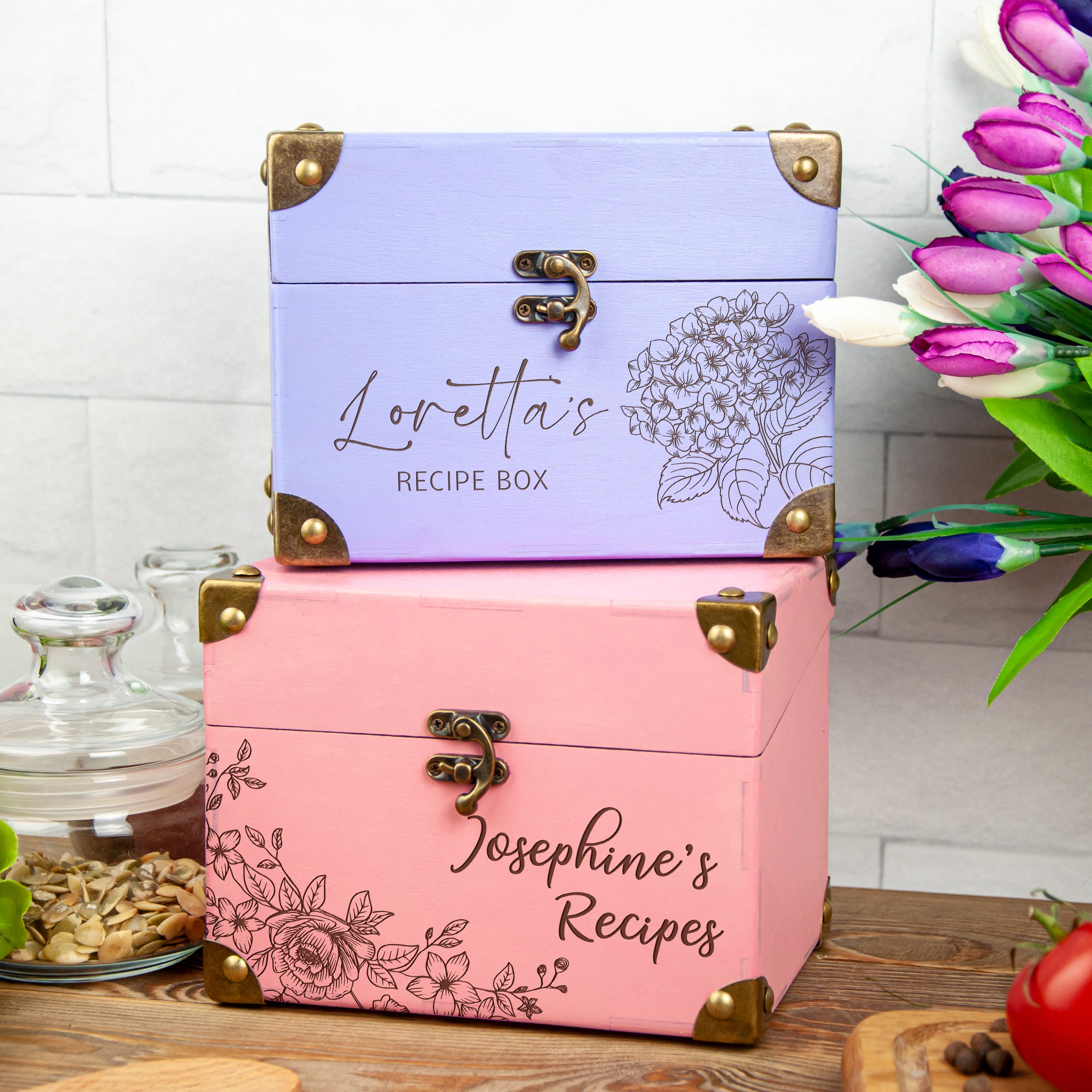 A pink and purple recipe box with the names "Loretta's RECIPE BOX" and "Josephine's Recipes" printed on it, along with a floral design. The boxes are placed on a wooden surface, with a vase of flowers and other decorative items in the background.