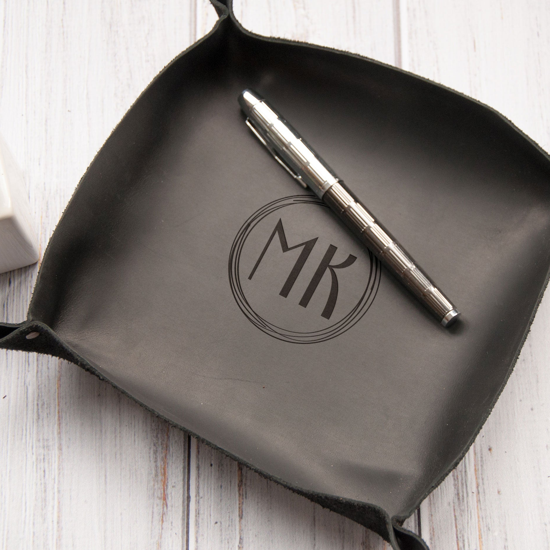 A black leather tray with a silver pen and a black circular logo on it, placed on a white wooden surface.