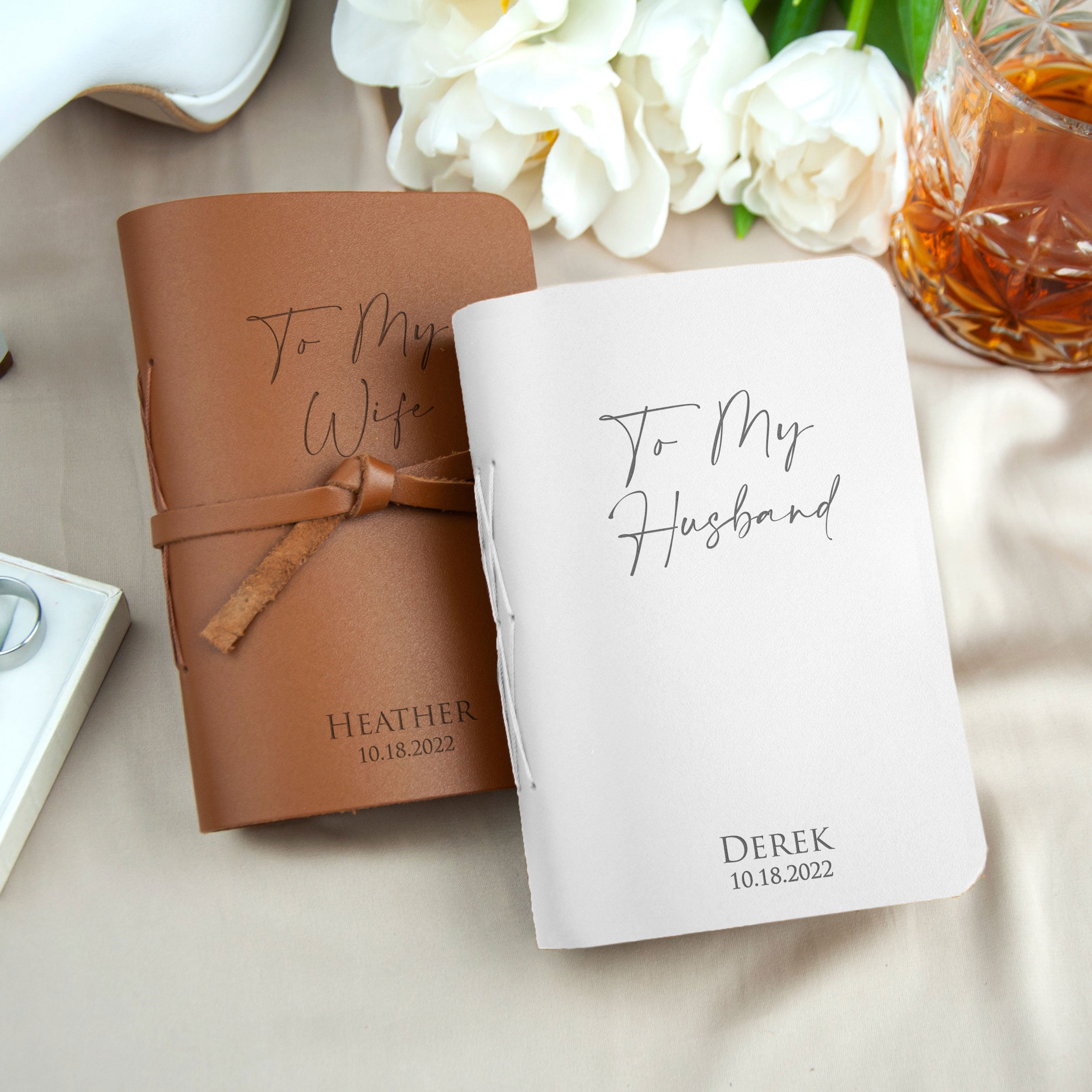 two personalized notebooks, one brown and one white, with the names "To My Wife" and "To My Husband" printed on them. The notebooks are placed on a table with a glass of wine and white flowers in the background.