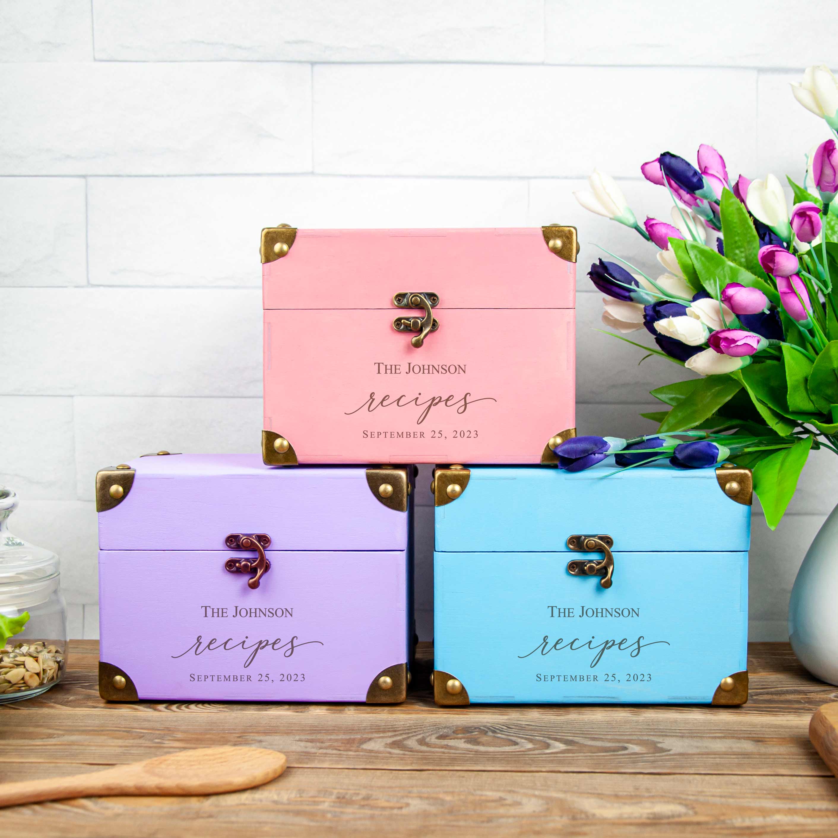 Three personalized recipe boxes with the name "The Johnson Recipes" printed on them, placed on a wooden surface. The boxes are in different colors - pink, blue, and purple. There is also a vase of flowers in the background.