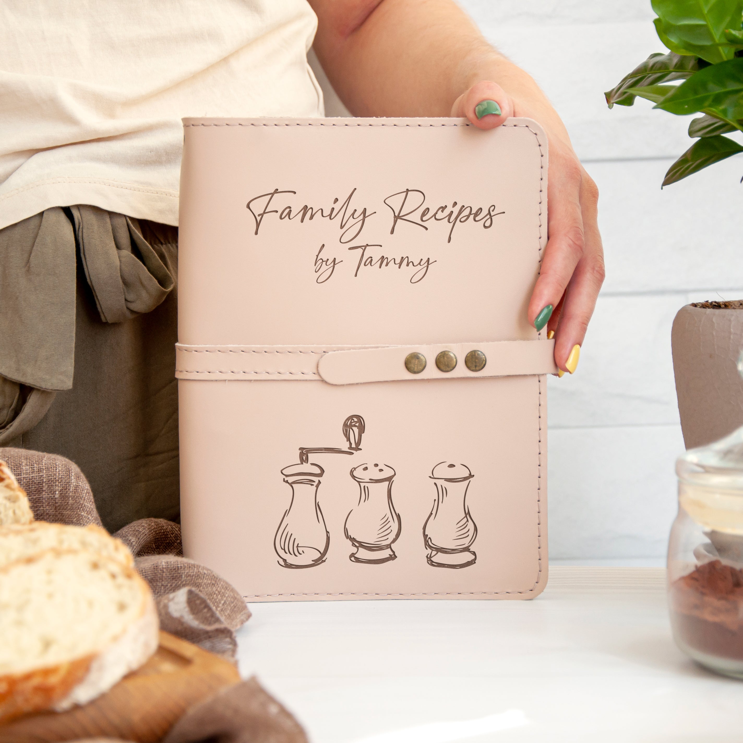 A person is holding a book titled "Family Recipes by Tanya" with a design of salt and pepper shakers on the cover.