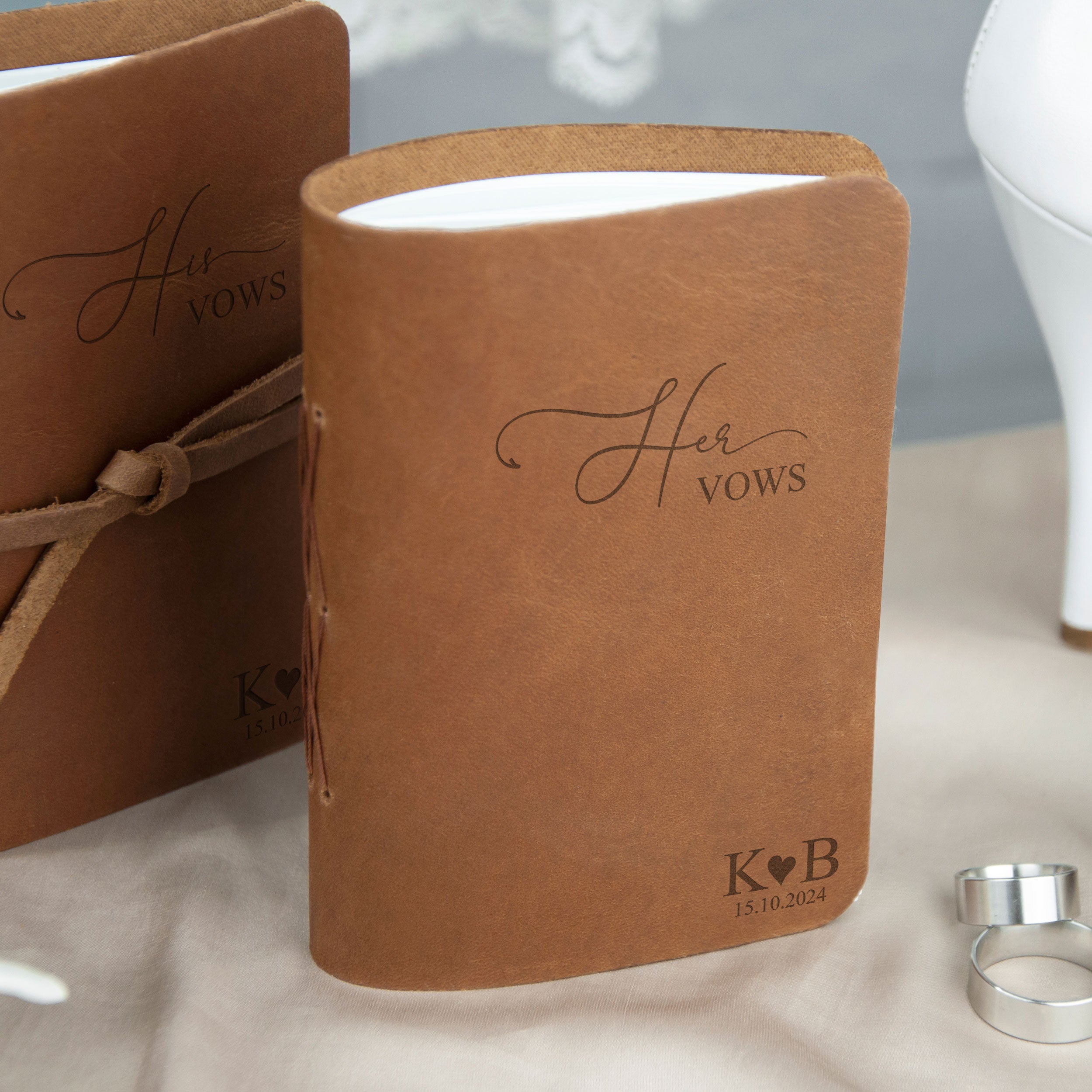 a leather-bound book titled "Her Vows" with a personalized message and date printed on it. The book is placed on a table with other items, such as rings, suggesting it may be a wedding-related item.
