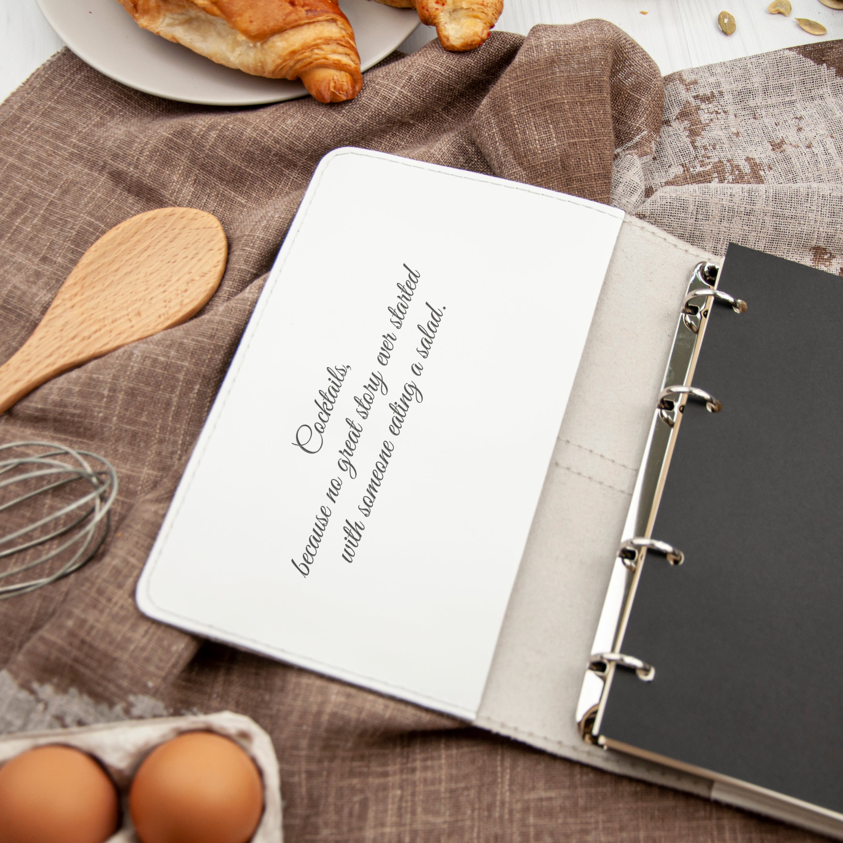a white notebook with a cursive text on it, placed on a table with various food items and kitchen utensils.