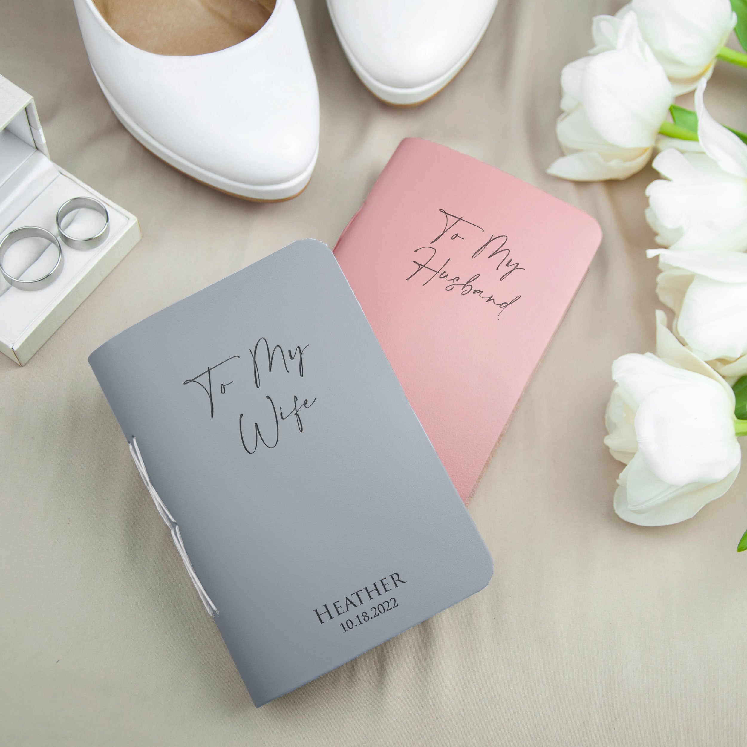 A gray notebook with the text "To My Wife" and a pink notebook with the text "To My Husband" are placed on a beige surface, along with a pair of white shoes and white flowers.