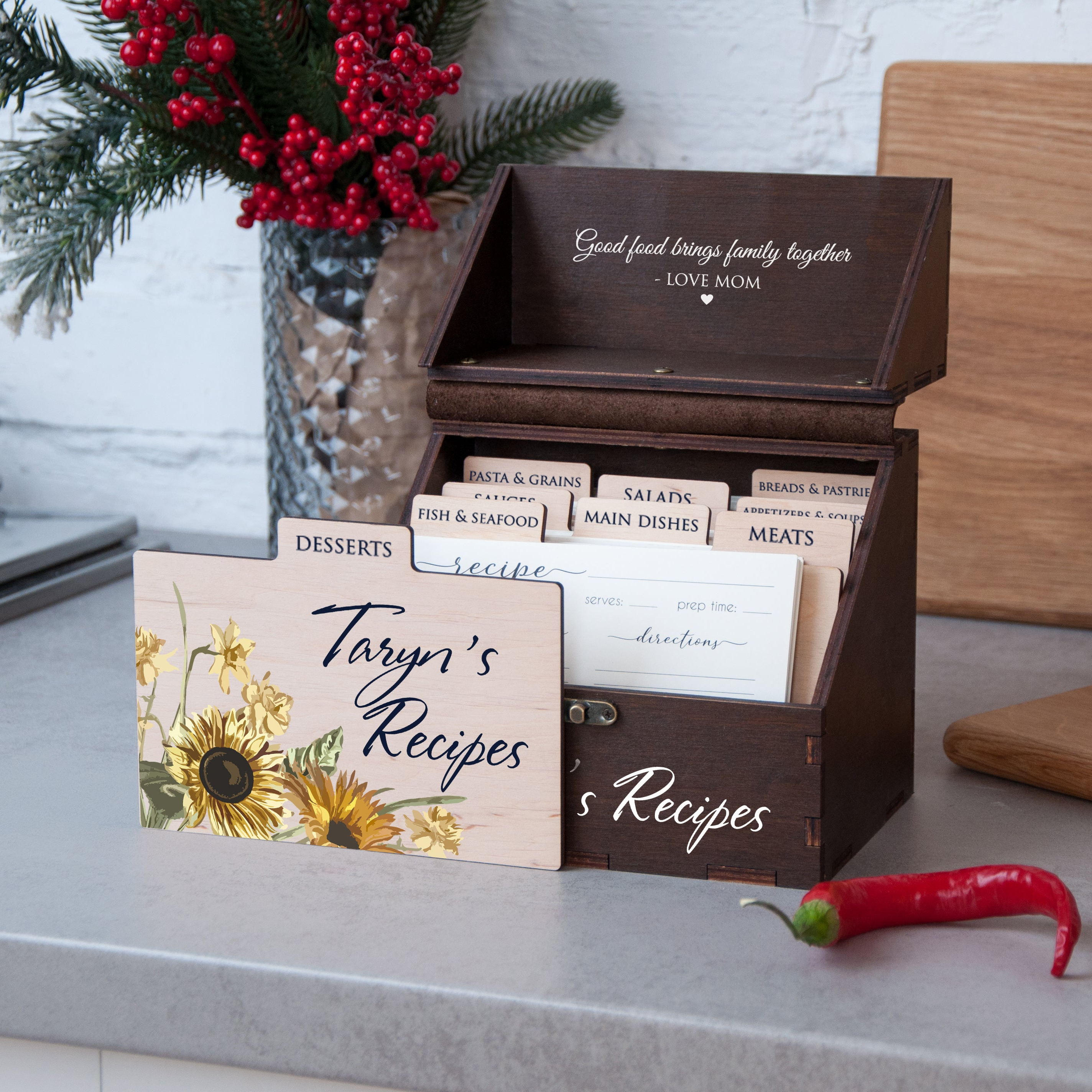 A wooden recipe box with a floral design, containing a collection of recipe cards, sits on a kitchen counter. The box is labeled "Taryn's Recipes" and has a vase of red berries and a wooden cutting board nearby.