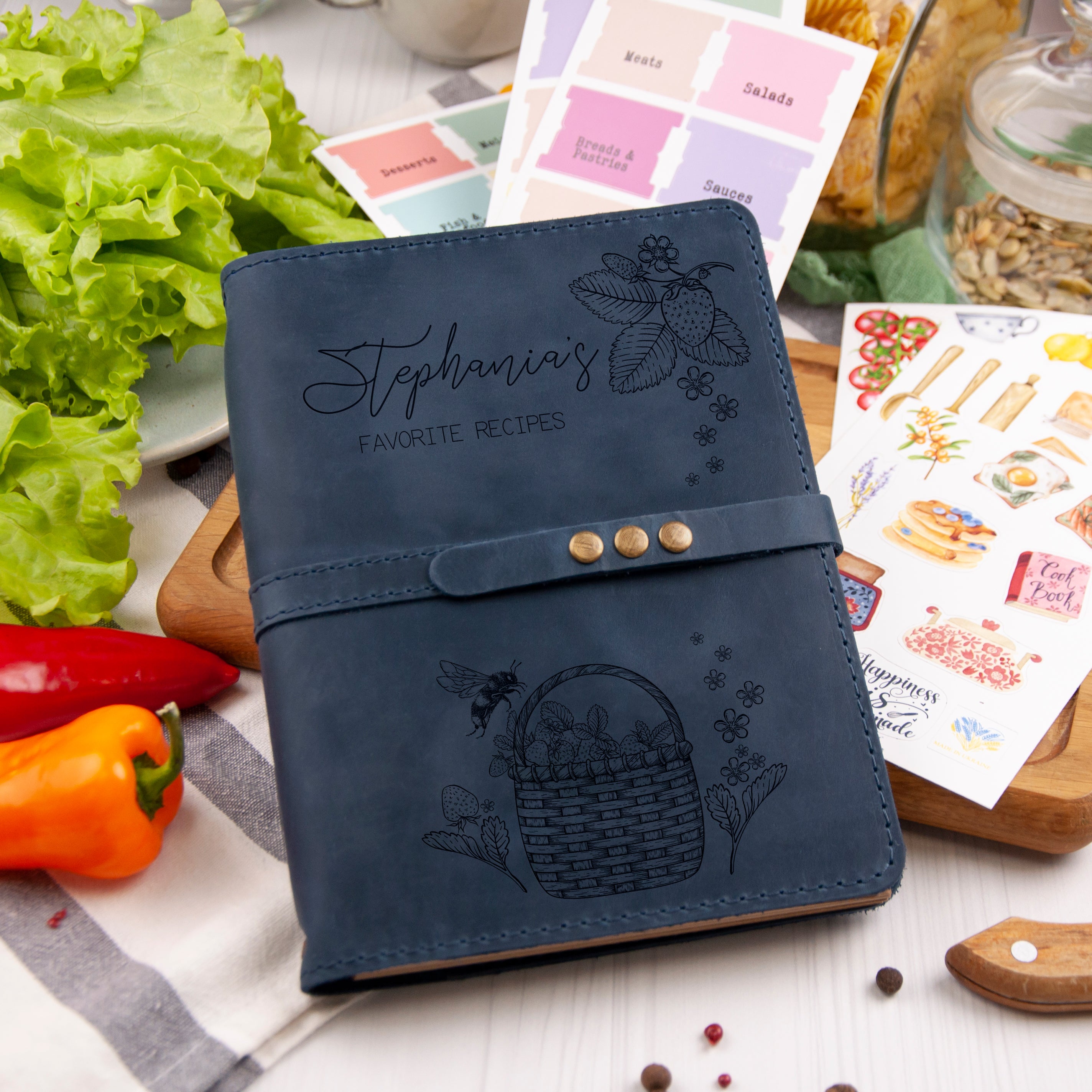 A blue leather-bound recipe book with a hand-drawn basket of produce sits on a wooden cutting board, surrounded by various kitchen items such as a red pepper, a green lettuce, and a jar of grains.