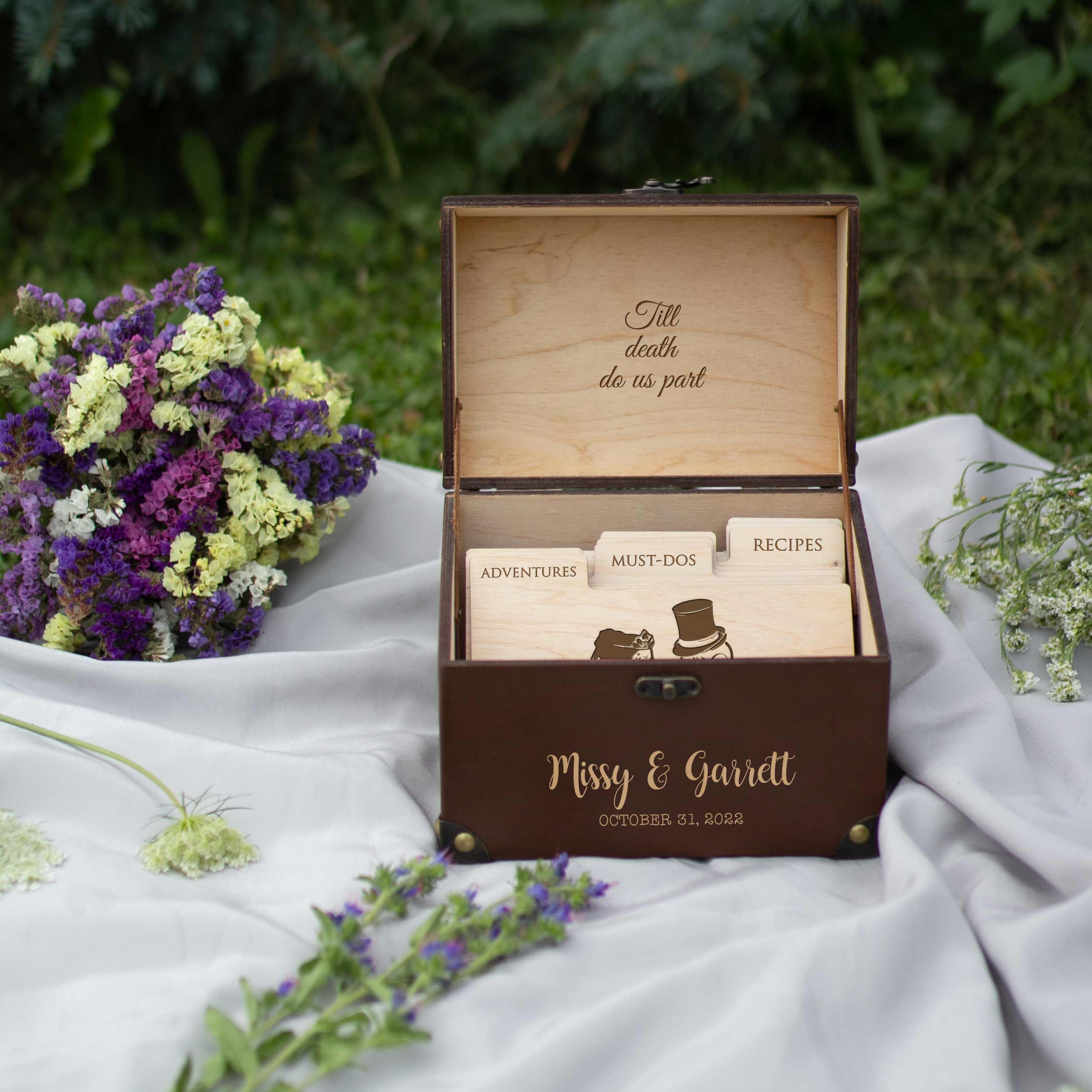 A wooden box with a lid, containing three wooden cards with illustrations and text, placed on a white cloth with purple and yellow flowers in the background.