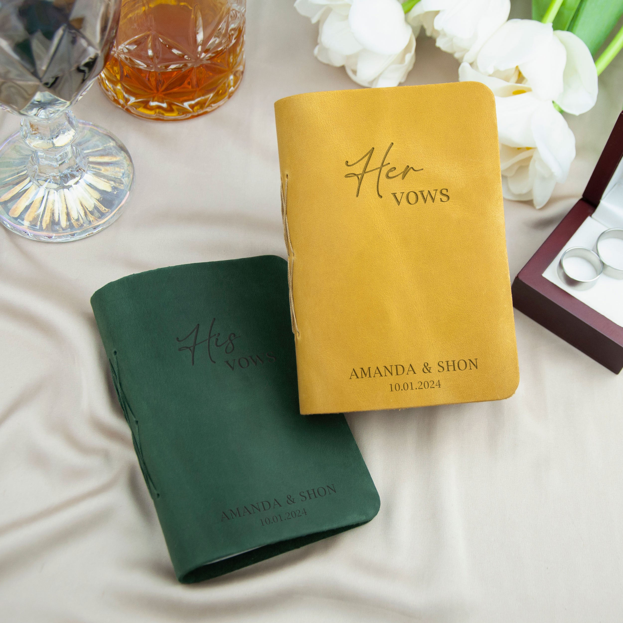 two personalized wedding vows books, one green and one yellow, placed on a table with a glass of wine and white flowers in the background.