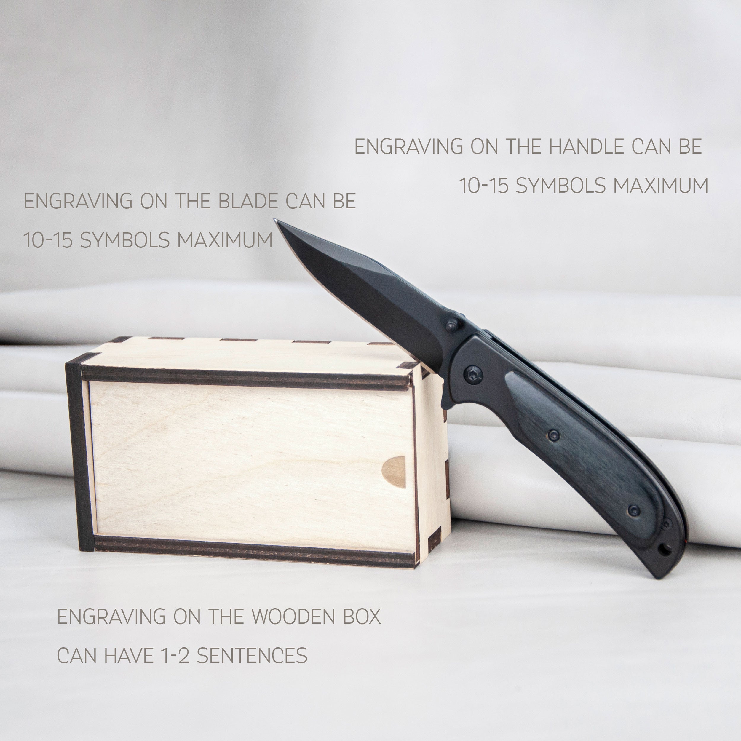 a black folding knife resting on a wooden box, with text above the objects stating that engraving on the handle can be up to 10-15 symbols maximum and that engraving on the wooden box can have 1-2 sentences.