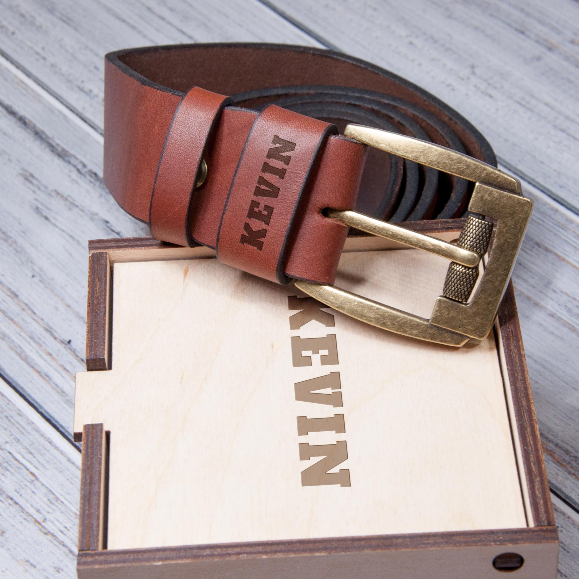 A brown leather belt with a personalized name engraved on it, placed on top of a wooden box with the name "KEVIN" printed on it.