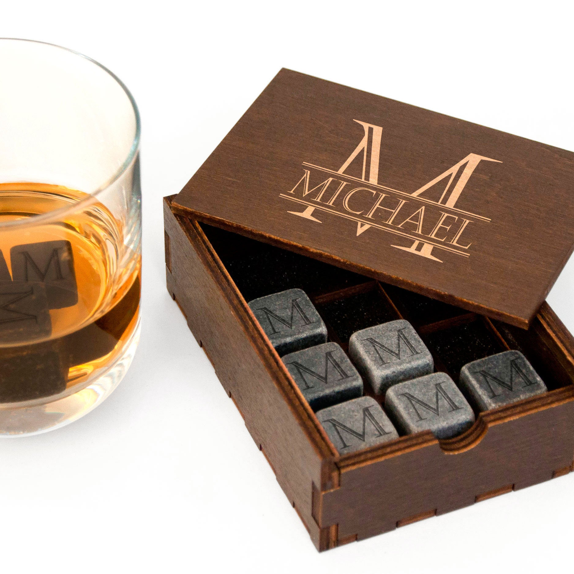 a wooden box with a lid, containing a set of gray marble slabs with the initials "M" engraved on them. Next to the box is a glass filled with a golden liquid, likely whiskey.