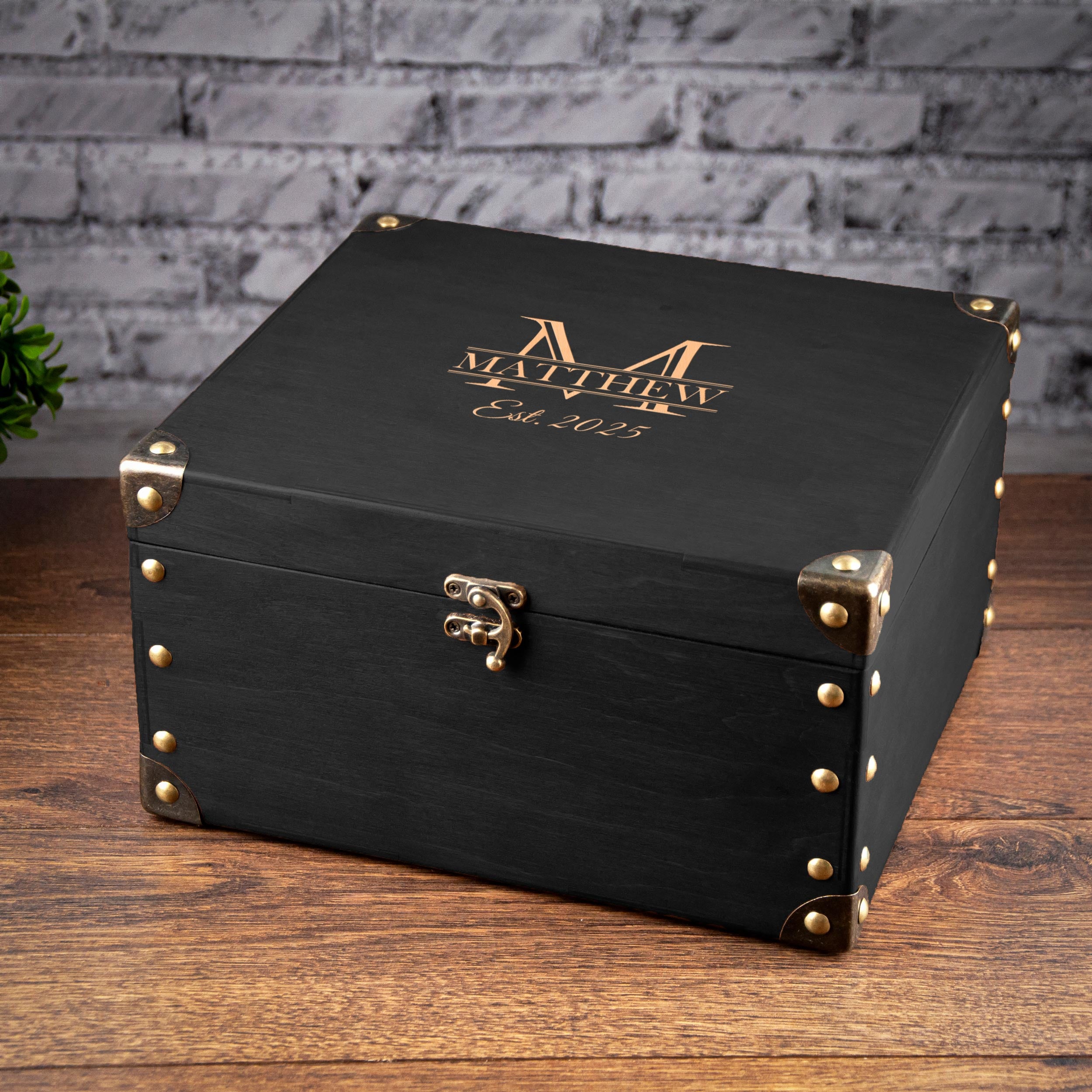 A black wooden box with gold-colored metal clasps and a gold-colored logo on the lid, placed on a wooden surface against a brick wall background.