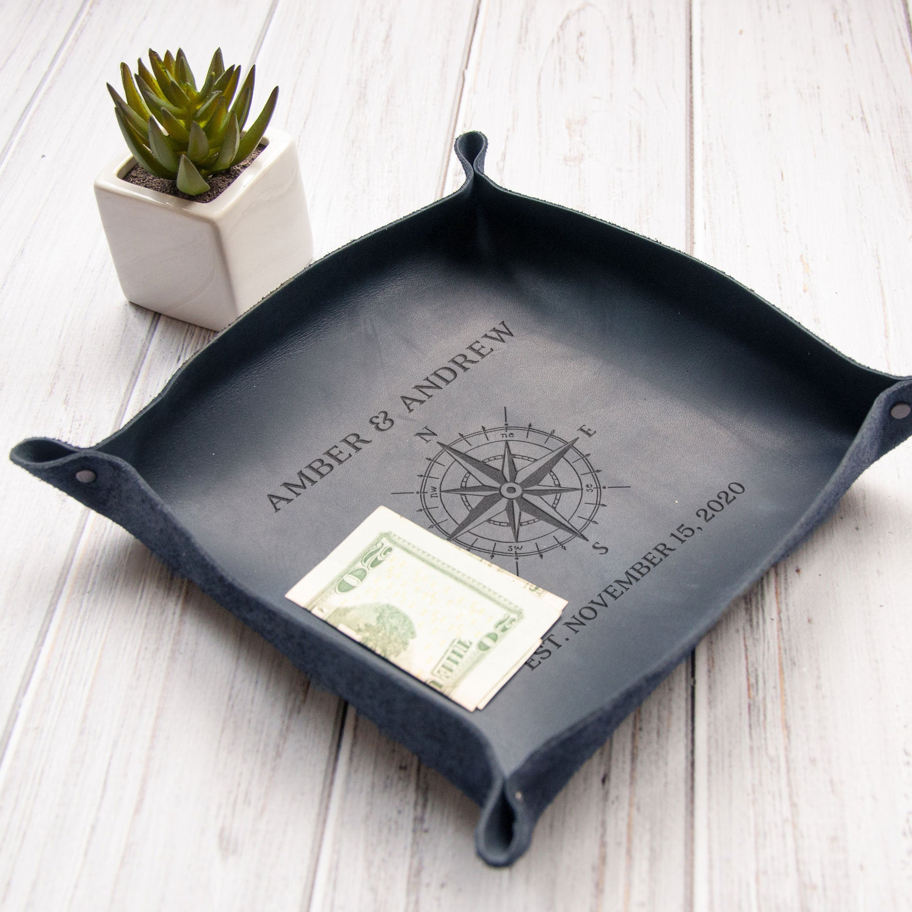 A black leather tray with a compass design and the names "AMBER & ANDREW" printed on it, along with the date "NOVEMBER 15, 2020". Next to the tray is a small potted succulent plant.