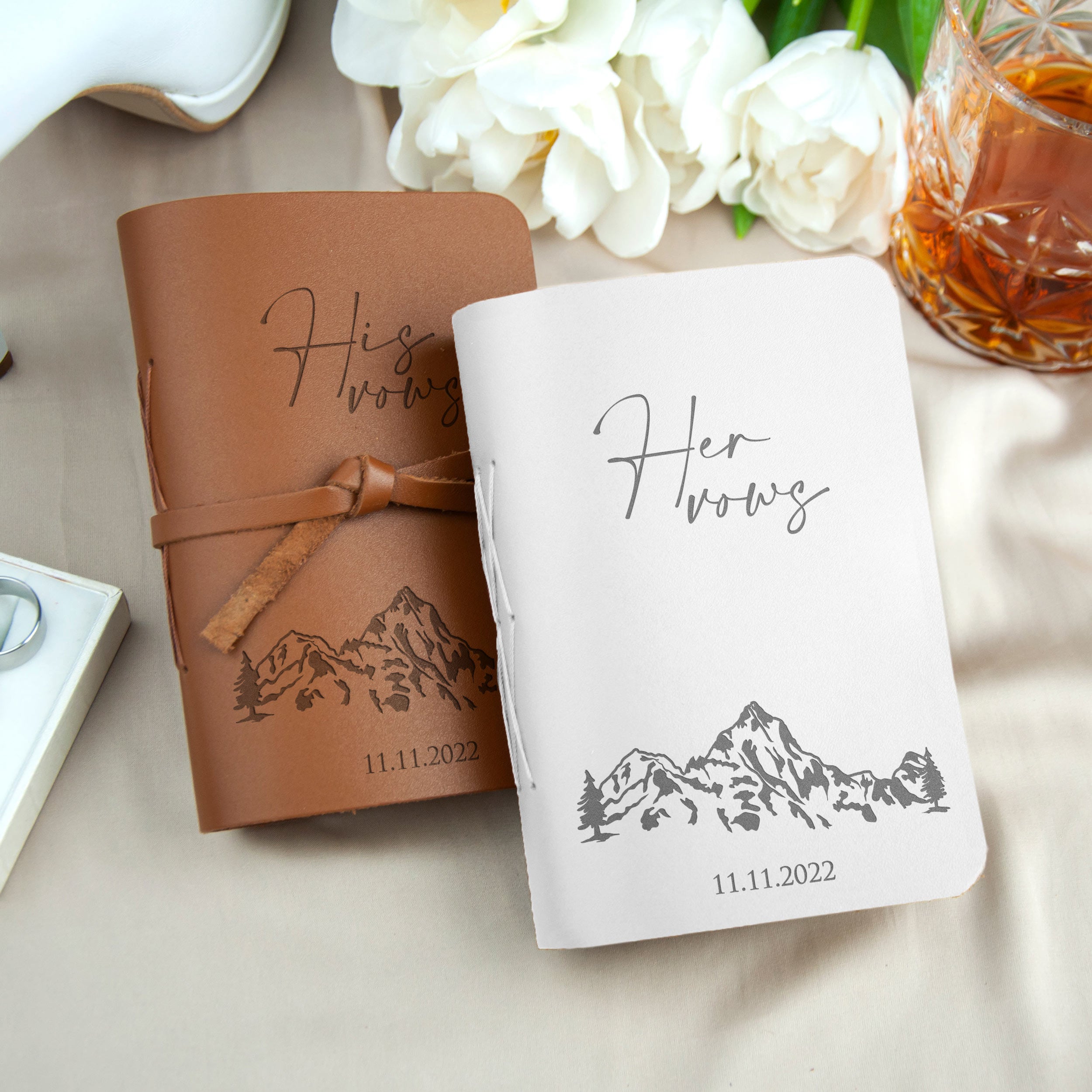 two leather-bound notebooks with the word "Her Vows" printed on them, placed on a table with a glass of what appears to be a beverage, and some flowers in the background.