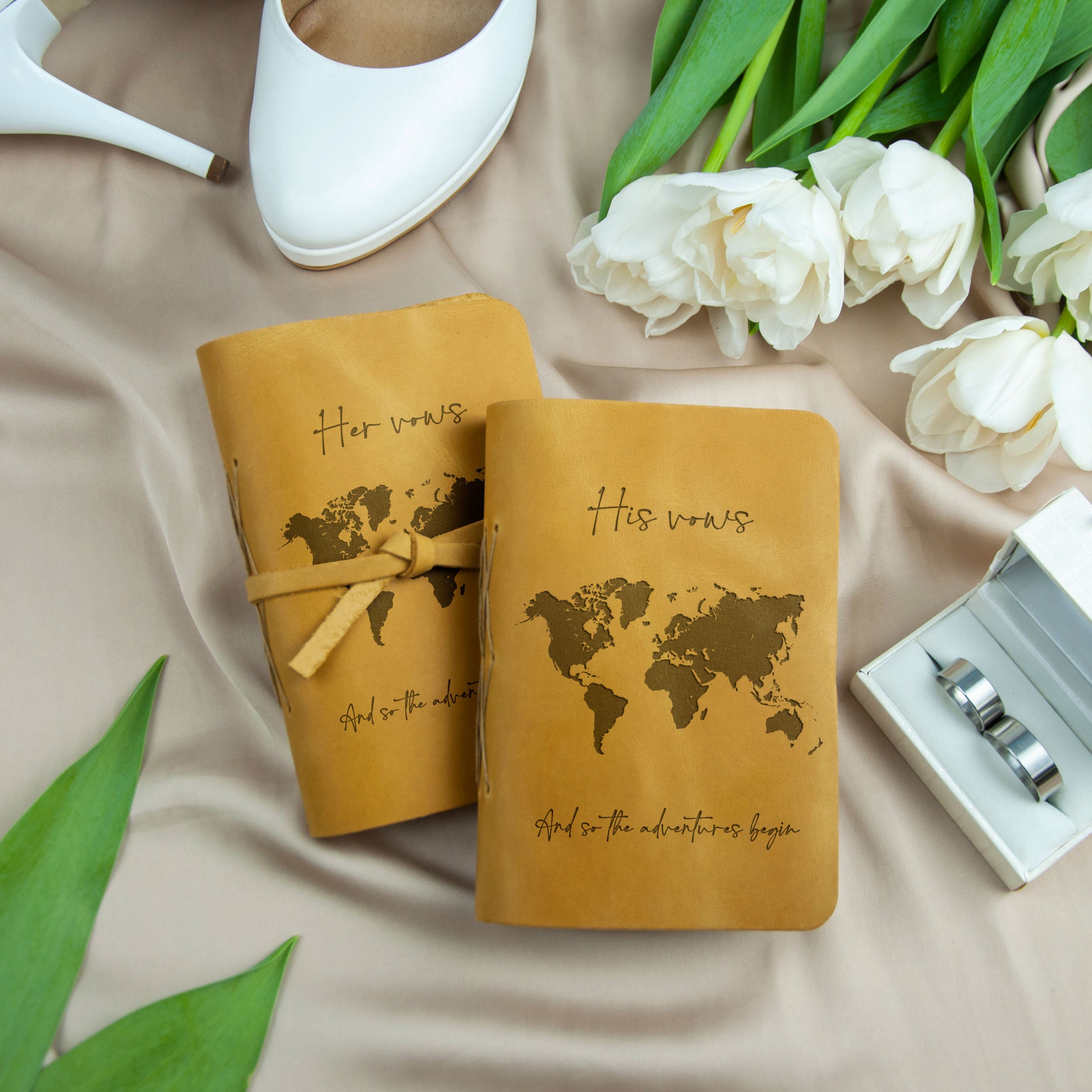a pair of leather-bound notebooks with a world map design, along with a pair of silver rings and a pair of white flowers.
