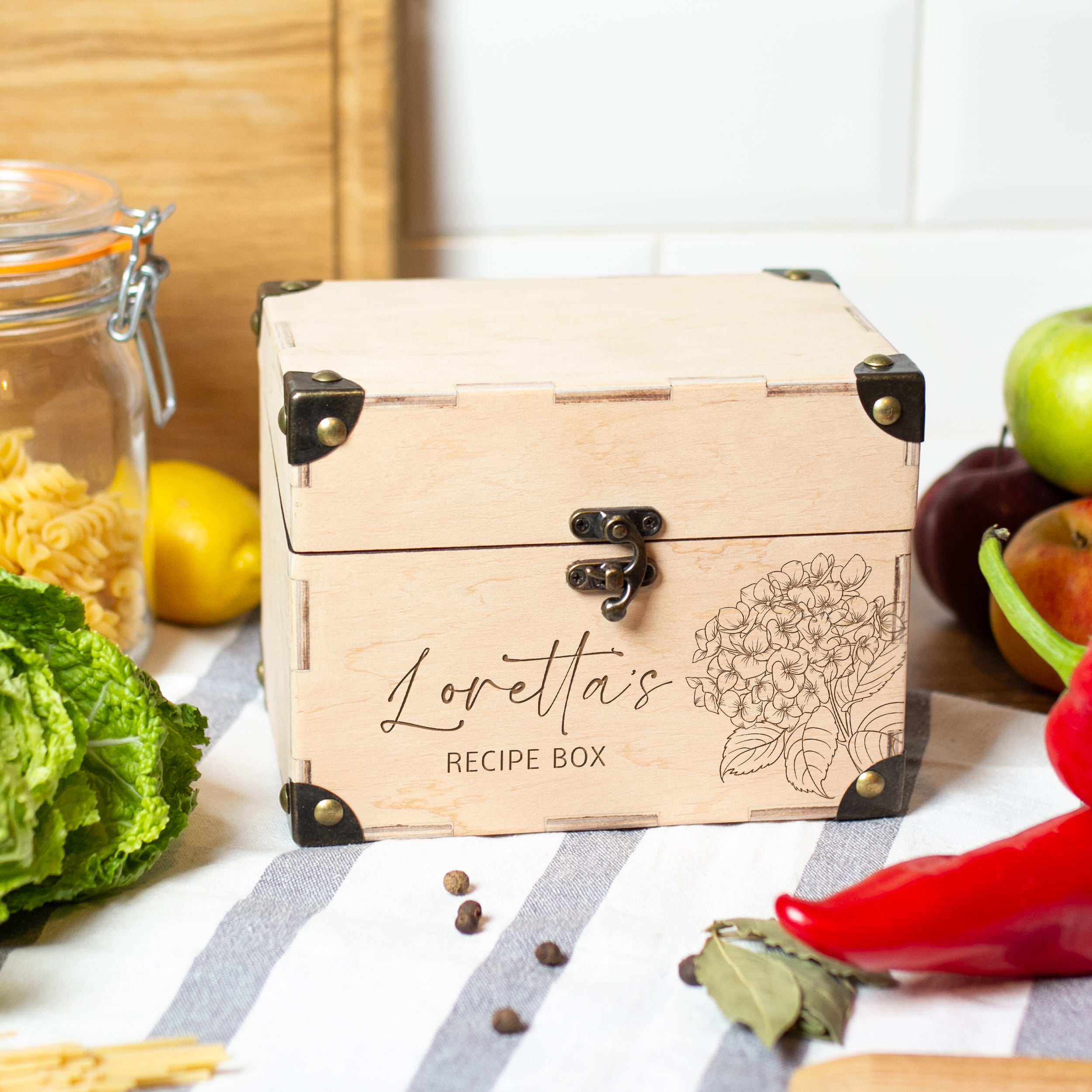 A wooden recipe box with a floral design, surrounded by various kitchen items such as a glass jar, apples, and peppers, on a kitchen counter.