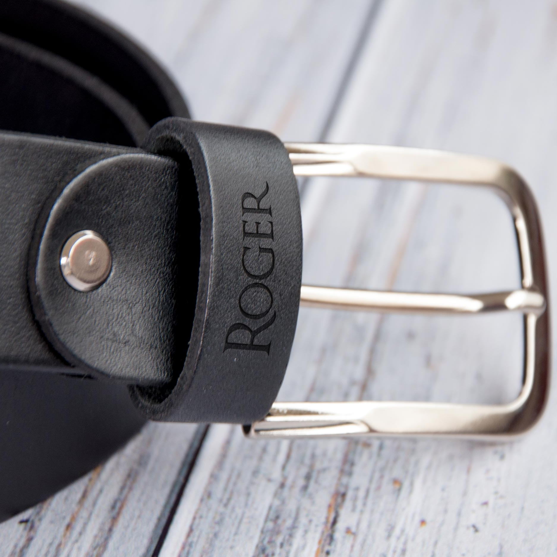 A close-up view of a black leather belt with a metal buckle, featuring the name "ROGER" engraved on the buckle.