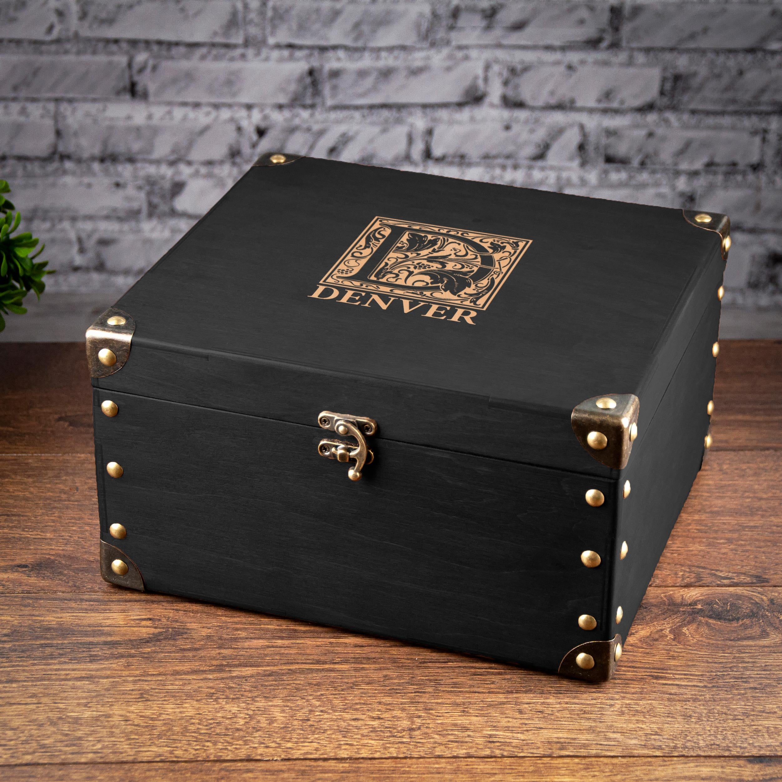 A black wooden box with a gold embossed logo, sitting on a wooden surface against a brick wall background.