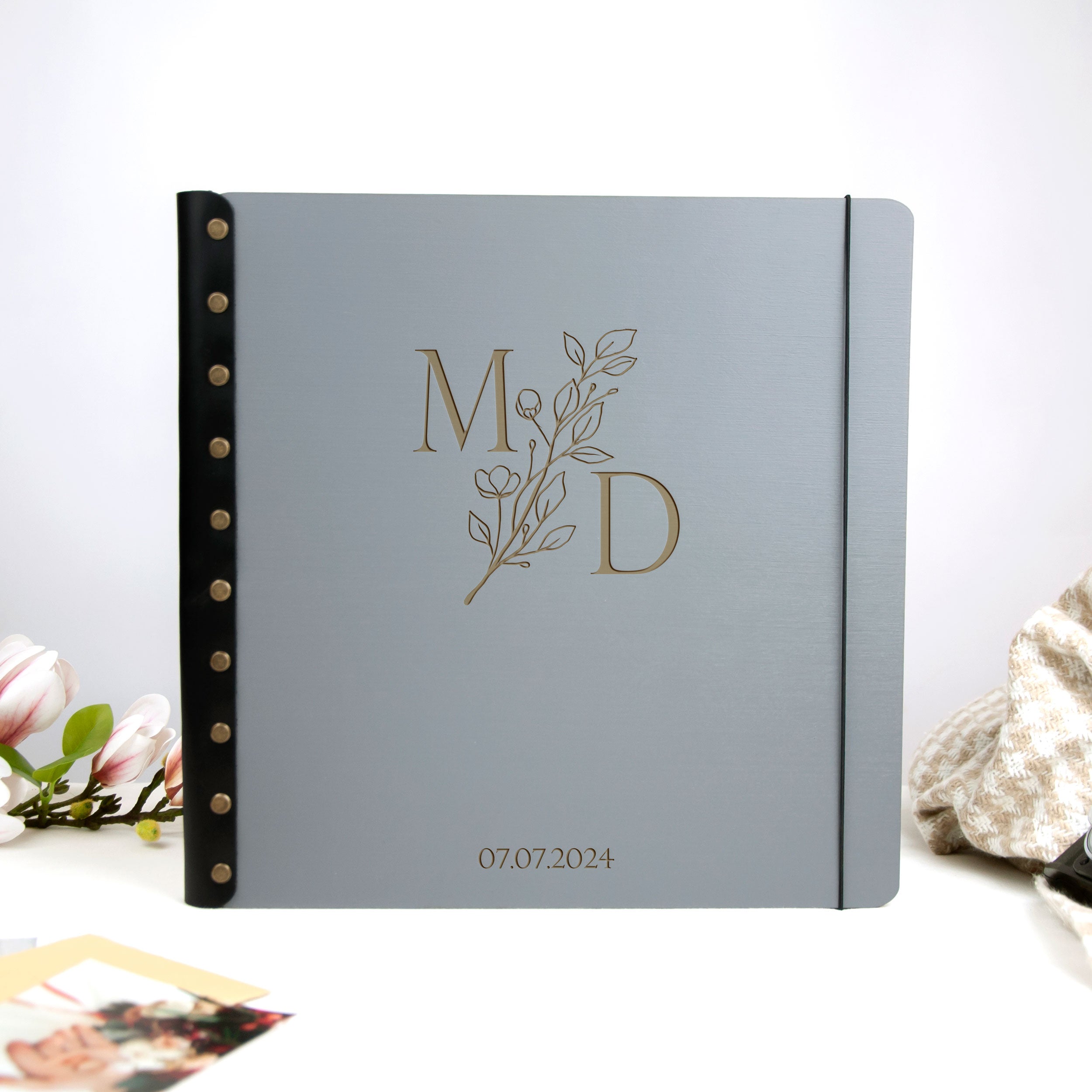 A gray photo album with a black leather spine and a gold monogrammed "M&D" logo, placed on a white surface with a floral arrangement in the background.
