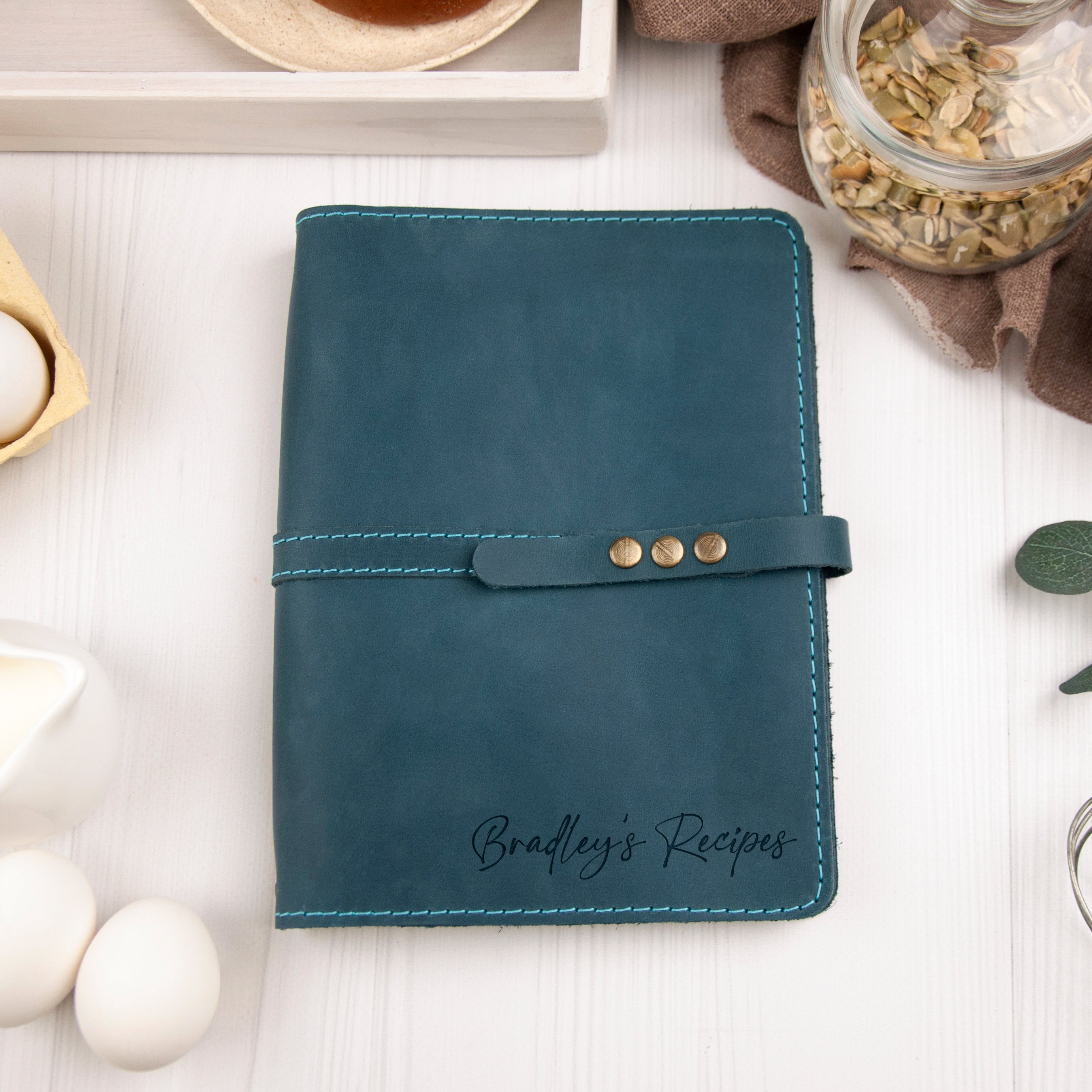 A teal leather recipe book with the name "Bradley's Recipes" printed on it, placed on a white surface alongside various kitchen items such as a jar of seeds, a white pitcher, and a white egg.