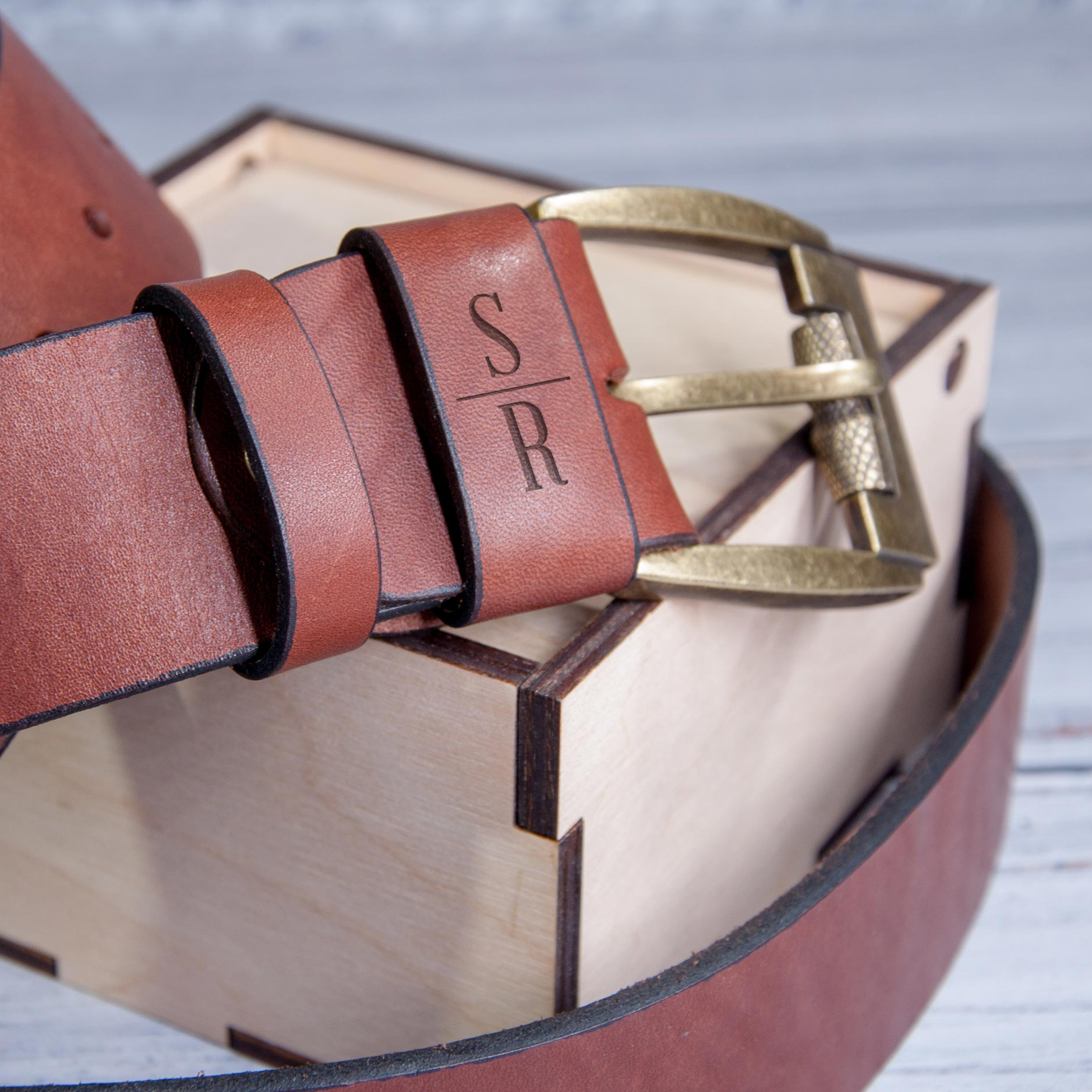 A brown leather belt with a personalized engraved buckle is placed on a wooden surface.