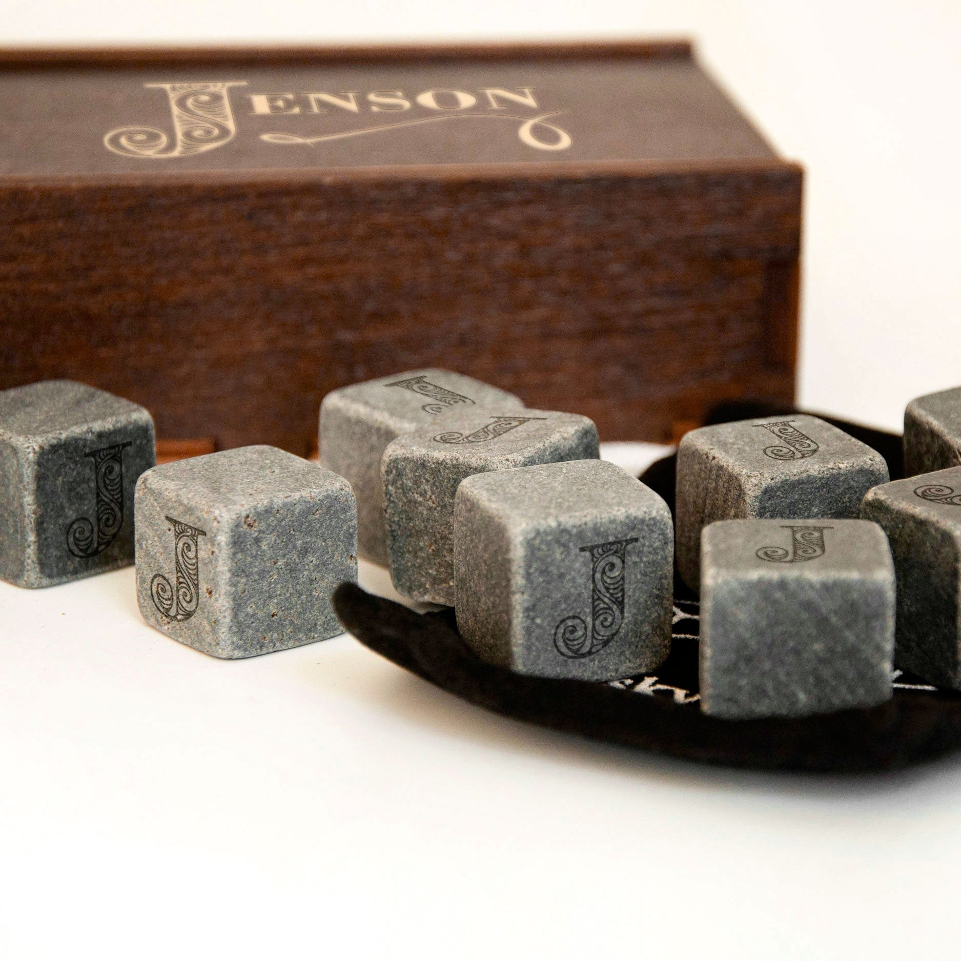 a wooden box with the word "Jenson" written on it, along with several gray stone cubes with the letter "J" engraved on them. The cubes are arranged on a black surface, with some of them placed on a black tray.