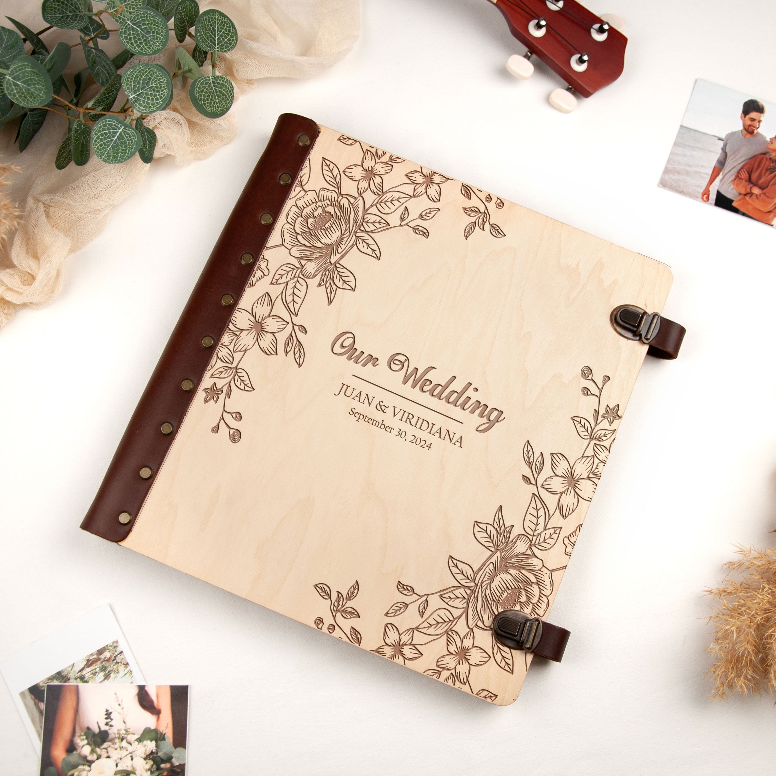 A wooden wedding album with a floral design, featuring the text "Our Wedding" and "Juan & Vridiana September 30, 2023". The album is placed on a white surface, surrounded by various items such as a guitar, a plant, and some photographs.