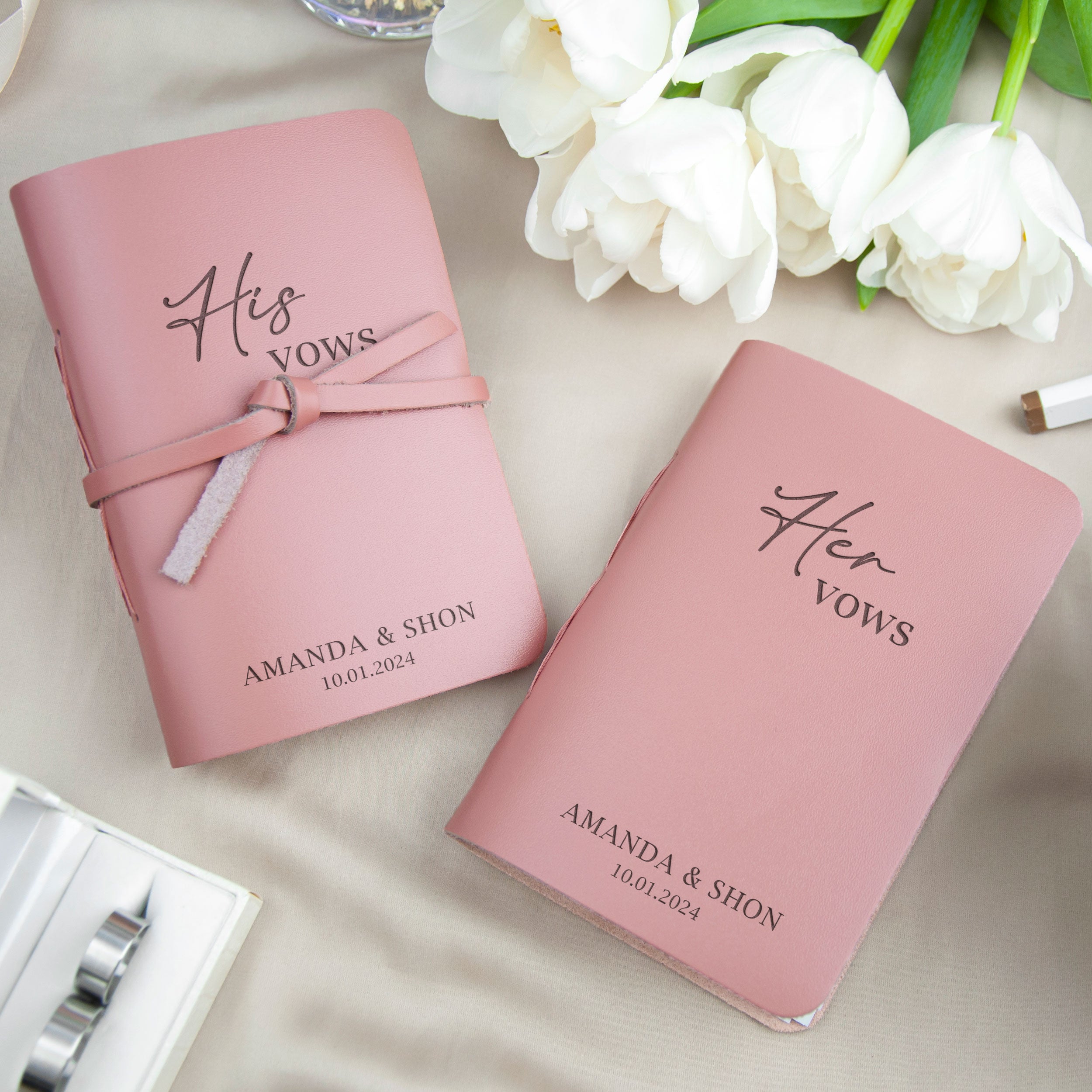 two pink notebooks with the text "His Vows" and "Her Vows" printed on them, along with the names "AMANDA & SHON" and the date "10.01.2024". The notebooks are placed on a table with white flowers, and there is a glass and a pen visible in the background.
