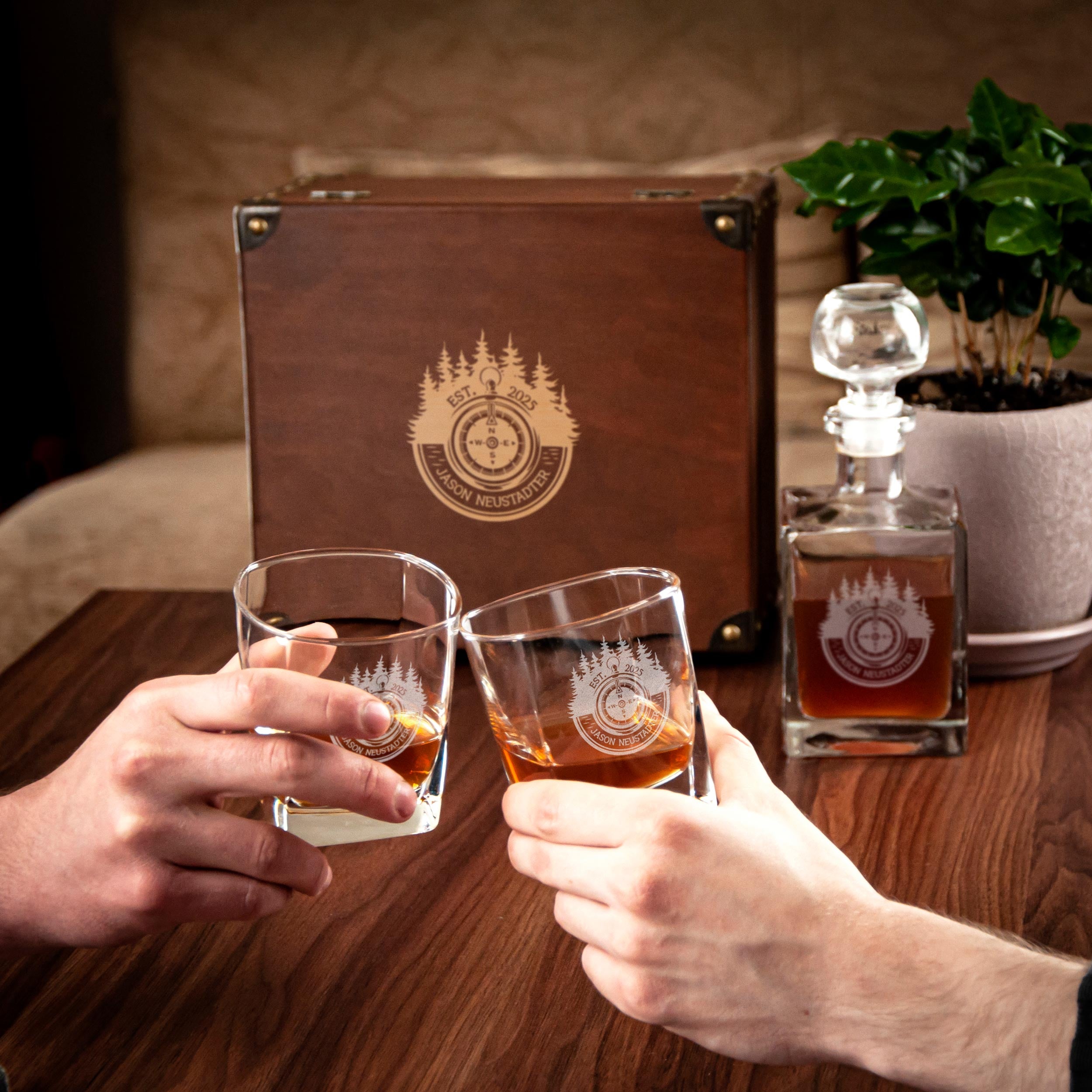 Two hands clink glasses of amber-colored liquid, likely whiskey, in front of a wooden box with a logo and a potted plant.