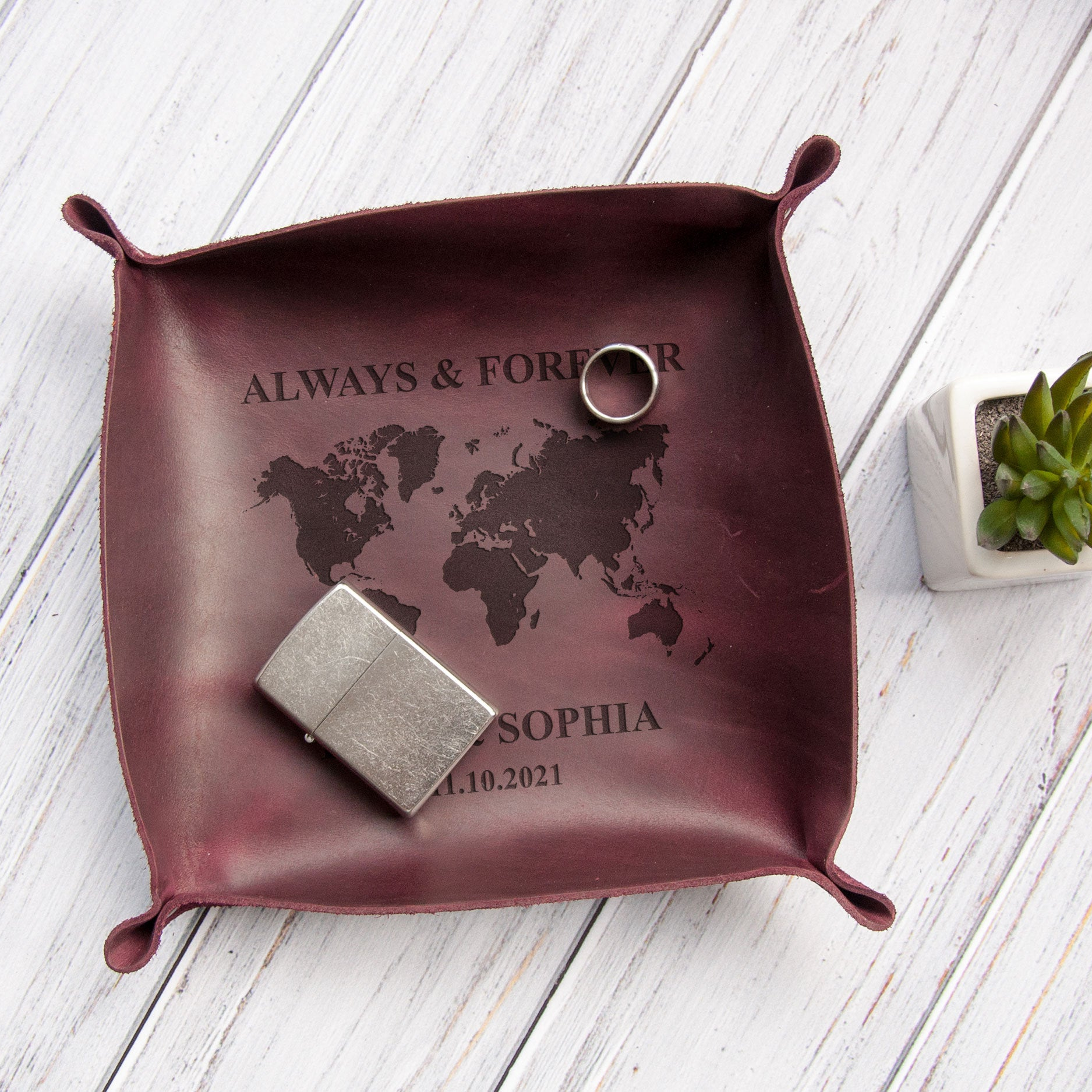 A maroon leather trinket tray with a world map design and the text "ALWAYS & FORVER" printed on it, along with a ring and a lighter, placed on a white wooden surface.