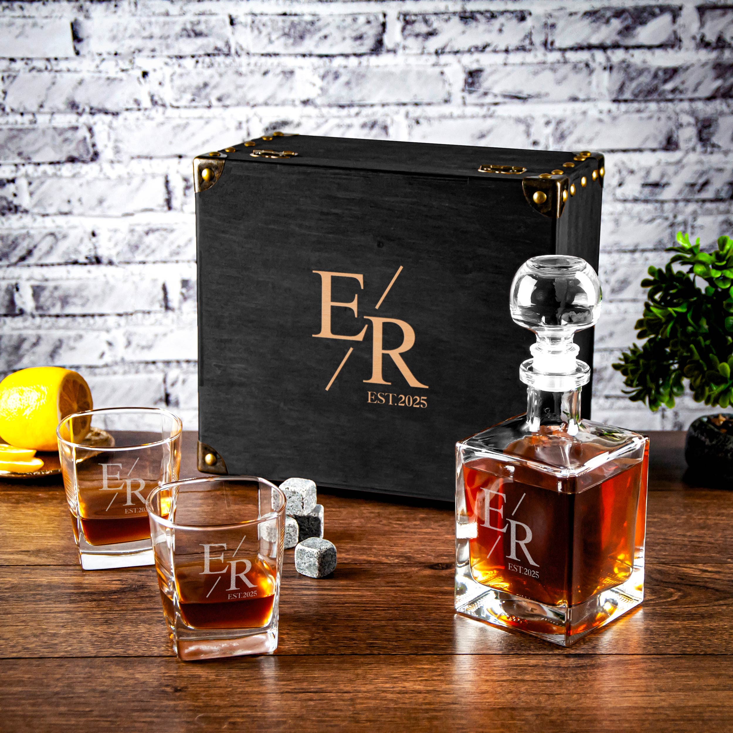 a set of three whiskey glasses and a bottle of whiskey, all placed on a wooden surface. The glasses are arranged in a row, with the bottle positioned next to them. In the background, there is a black box with the initials "ER" and the year "EST. 2015" printed on it.
