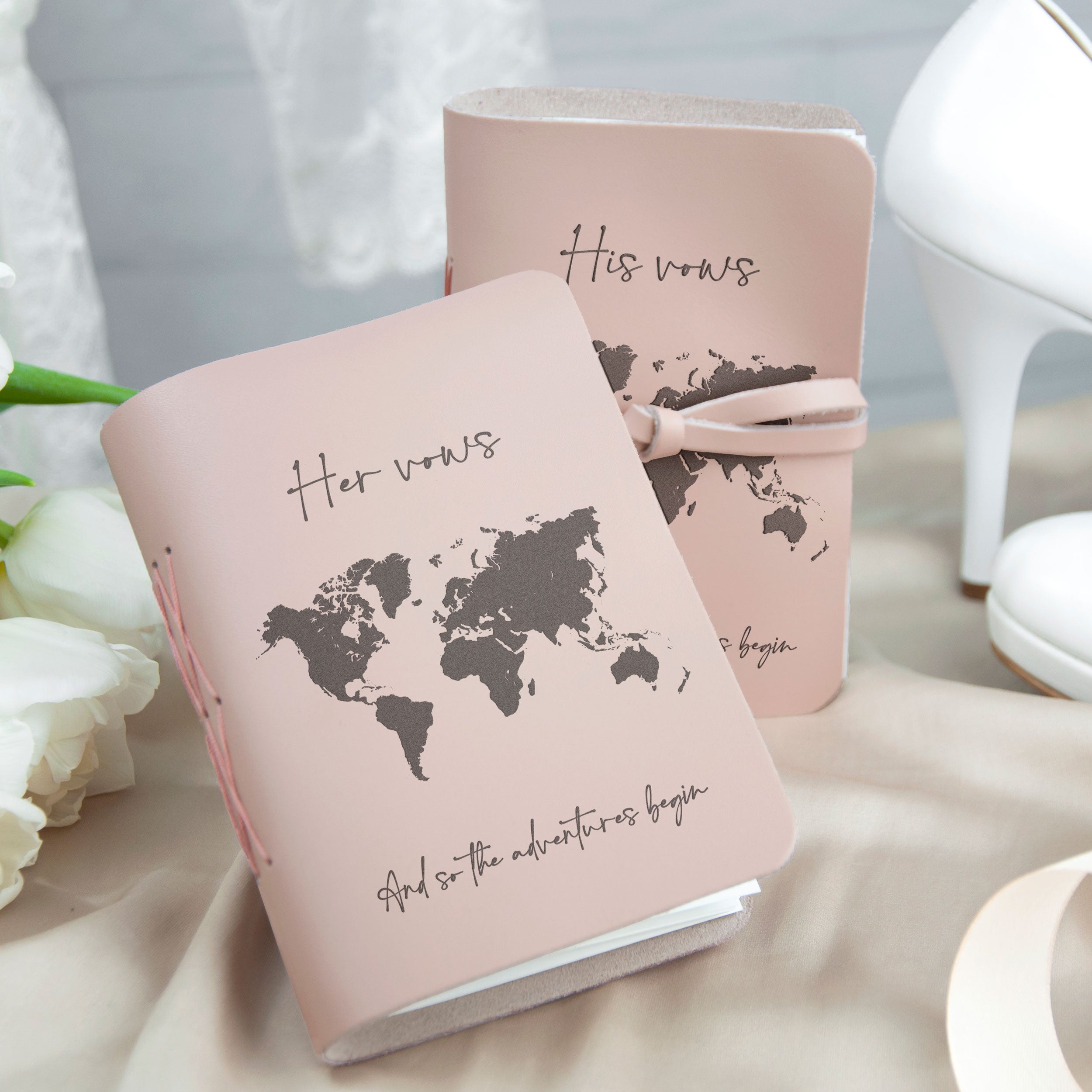 two pink notebooks with a world map design, one titled "Her vows" and the other titled "His vows". The notebooks are placed on a table with a white flower arrangement in the background.