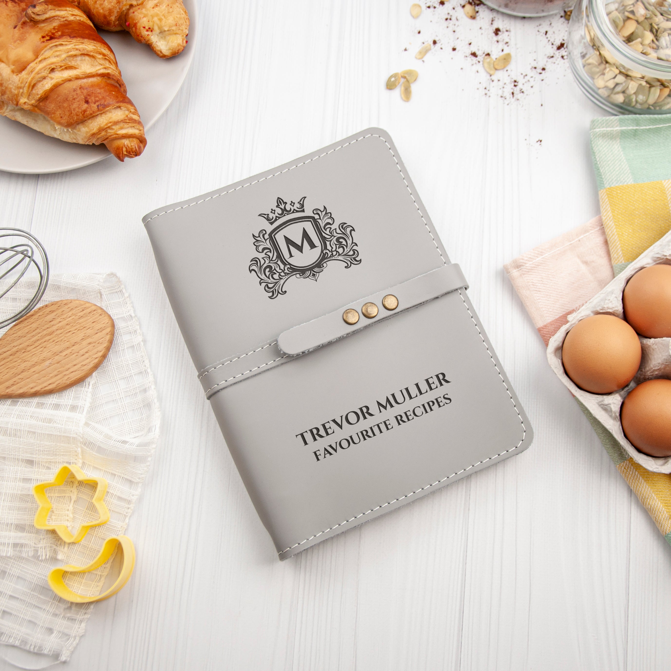 A personalized leather-bound recipe book with the name Trevor Muller' printed on the cover, along with the title 'Favourite Recipes', sits on a white surface surrounded by various kitchen items such as a croissant, eggs, and a wooden spoon.