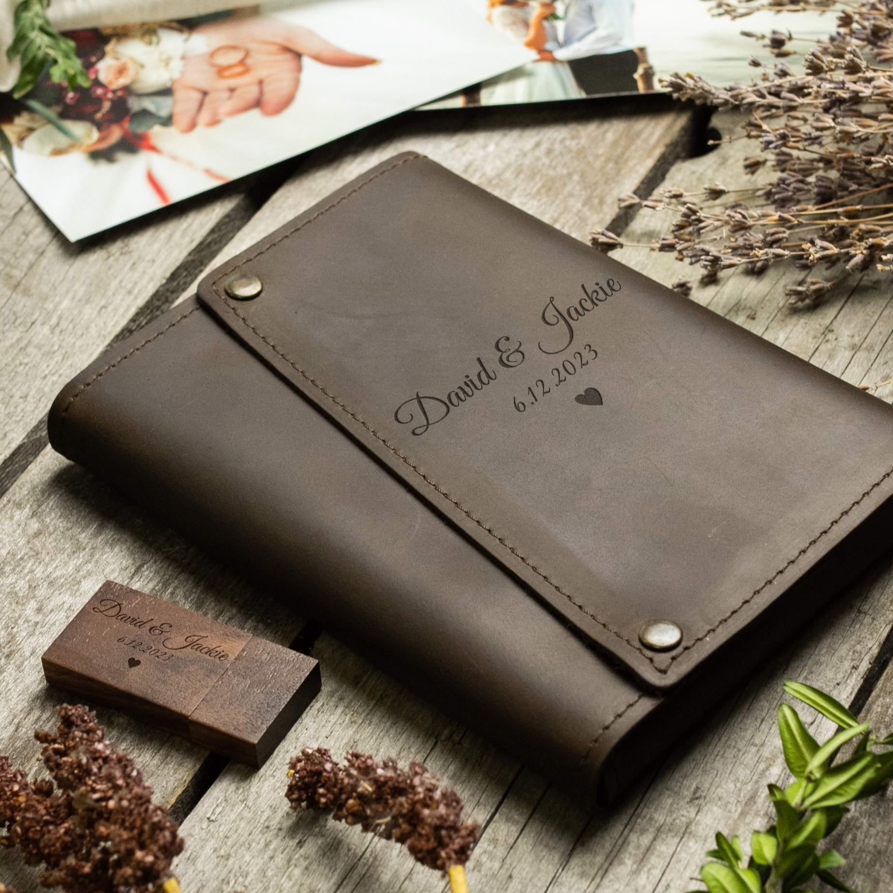 A leather-bound notebook with the names 'David & Jackie' and the date '6.12.2023' printed on it, along with a wooden block with the text 'David & Jackie' and the date '6.12.2023' printed on it. The notebook and wooden block are placed on a wooden surface, with some dried flowers and a photograph visible in the background.