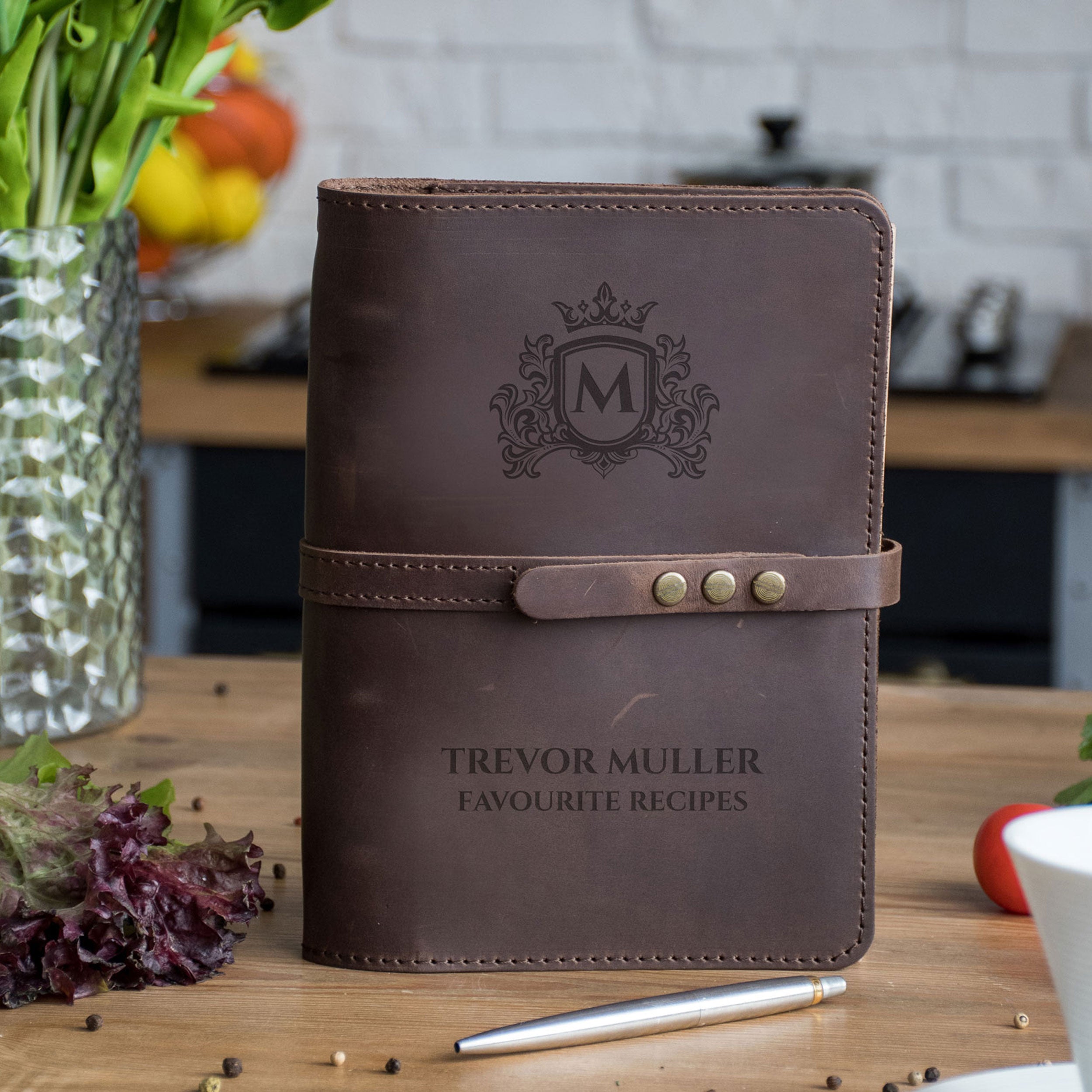 A brown leather recipe book with a monogrammed "M" logo, sitting on a wooden table with a vase of flowers and various kitchen items in the background.